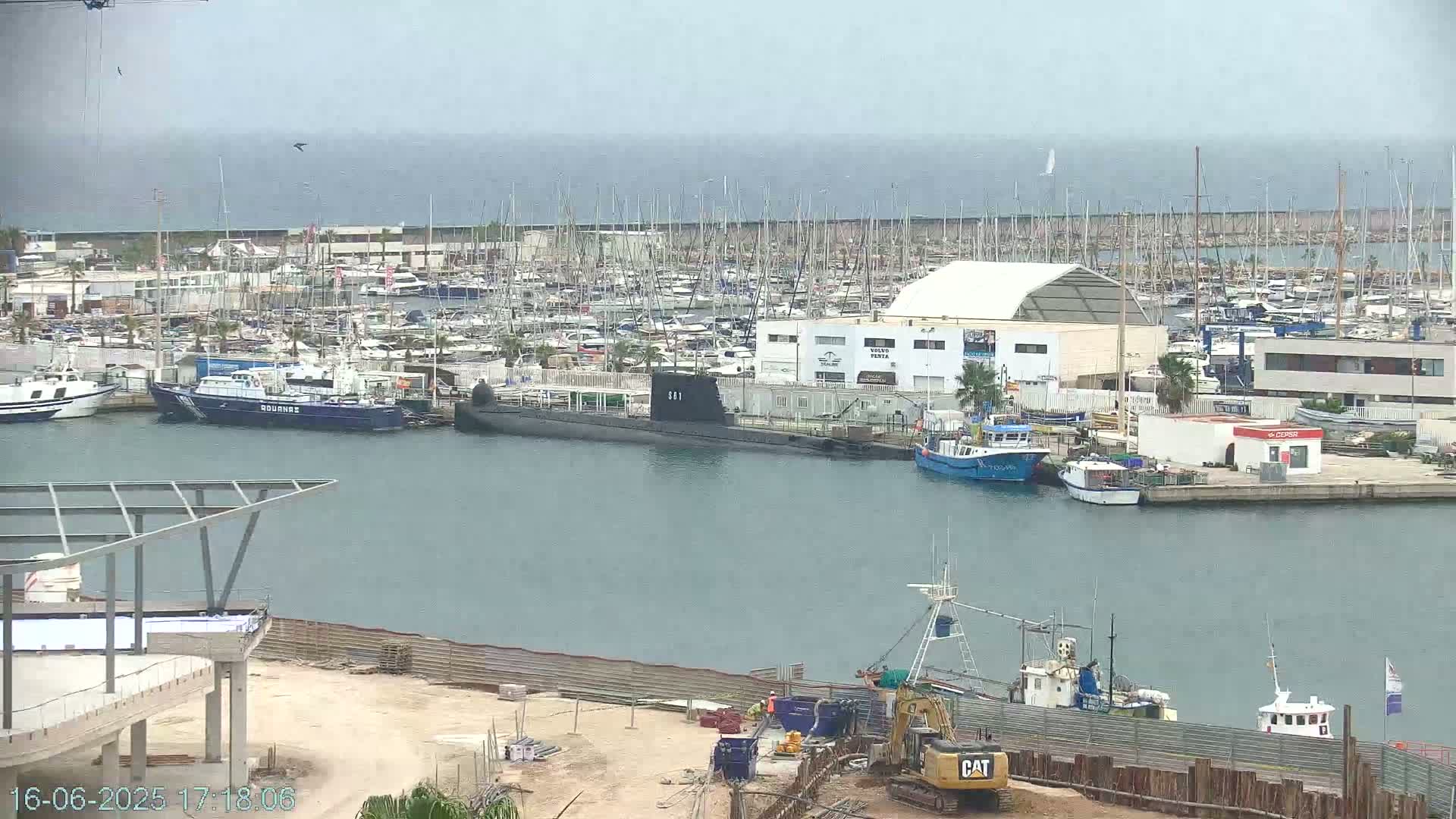 A marina filled with numerous boats and a docked submarine is seen under an overcast sky, with ongoing construction visible in the foreground.