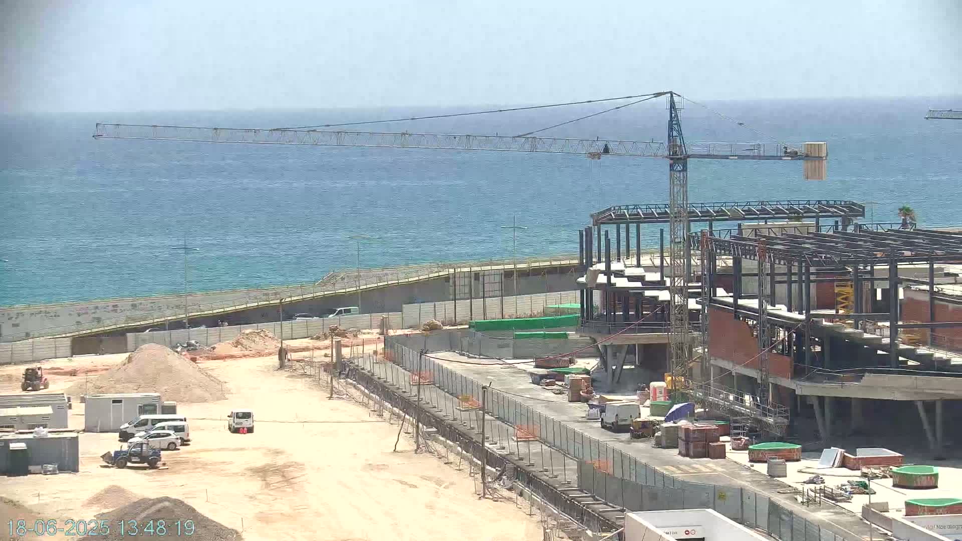 A coastal construction site with a large crane overlooks a calm ocean under a clear sky.