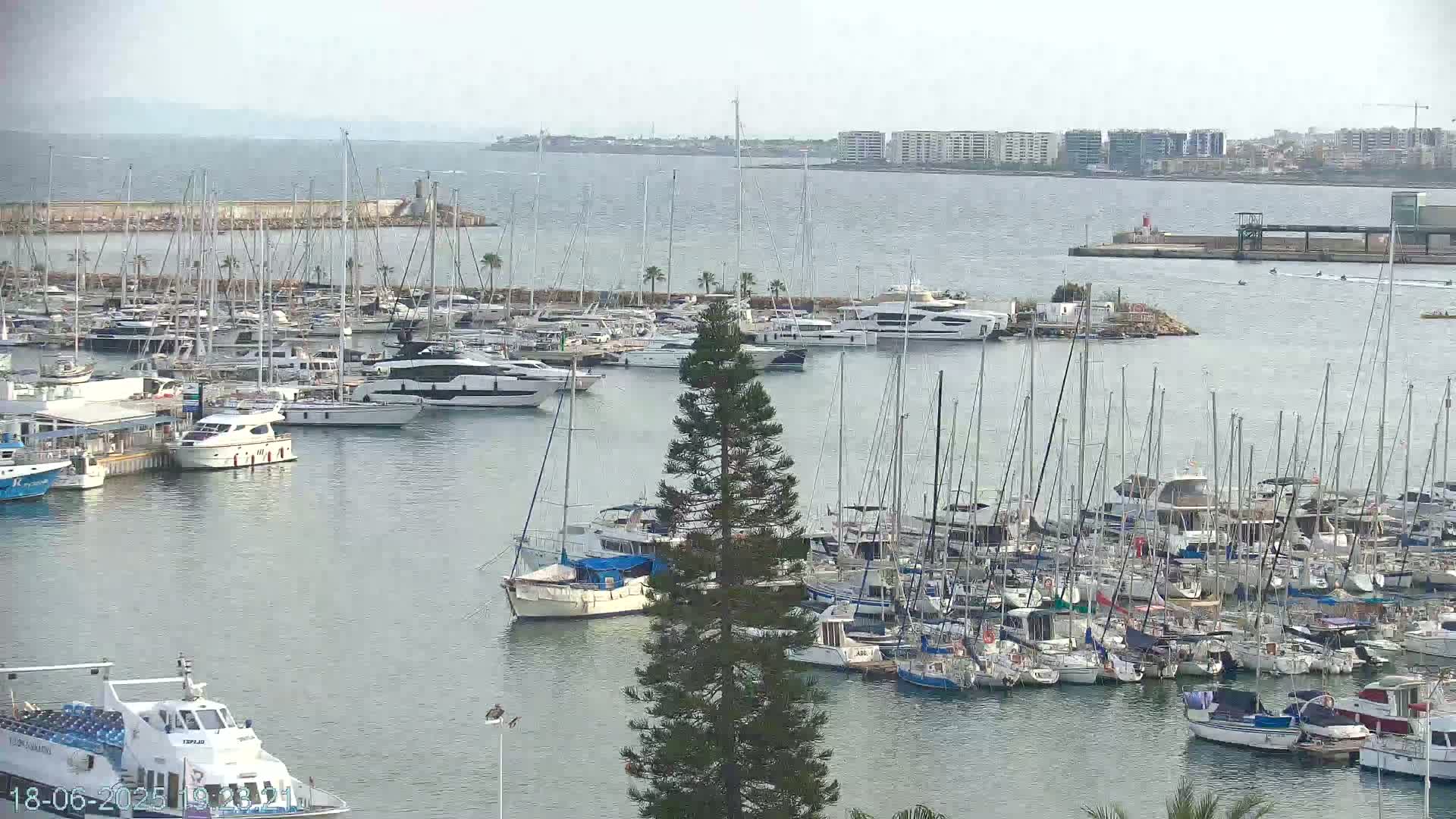 A marina filled with numerous sailboats and motorboats is seen on a calm, overcast day, with a distant cityscape visible across the water.