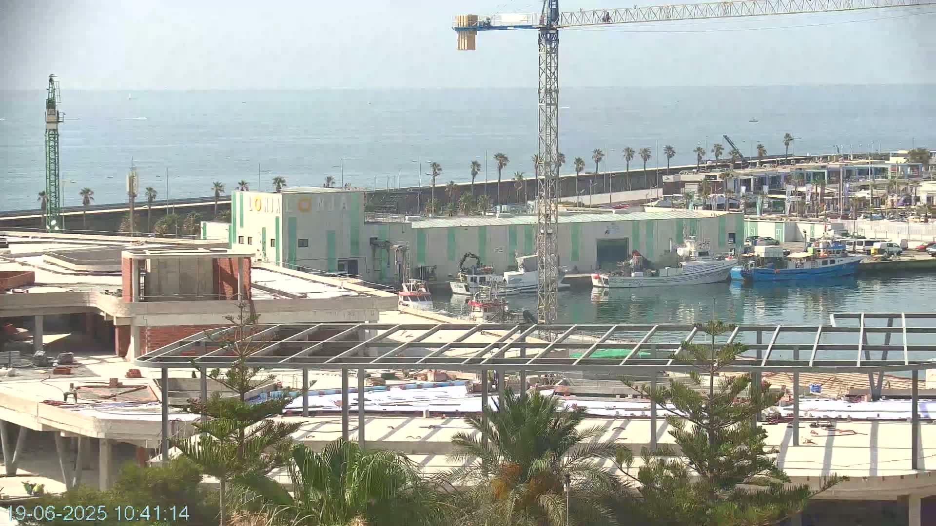 A partially constructed building sits near a harbor where several fishing boats are docked, under a mostly clear sky next to a calm sea.