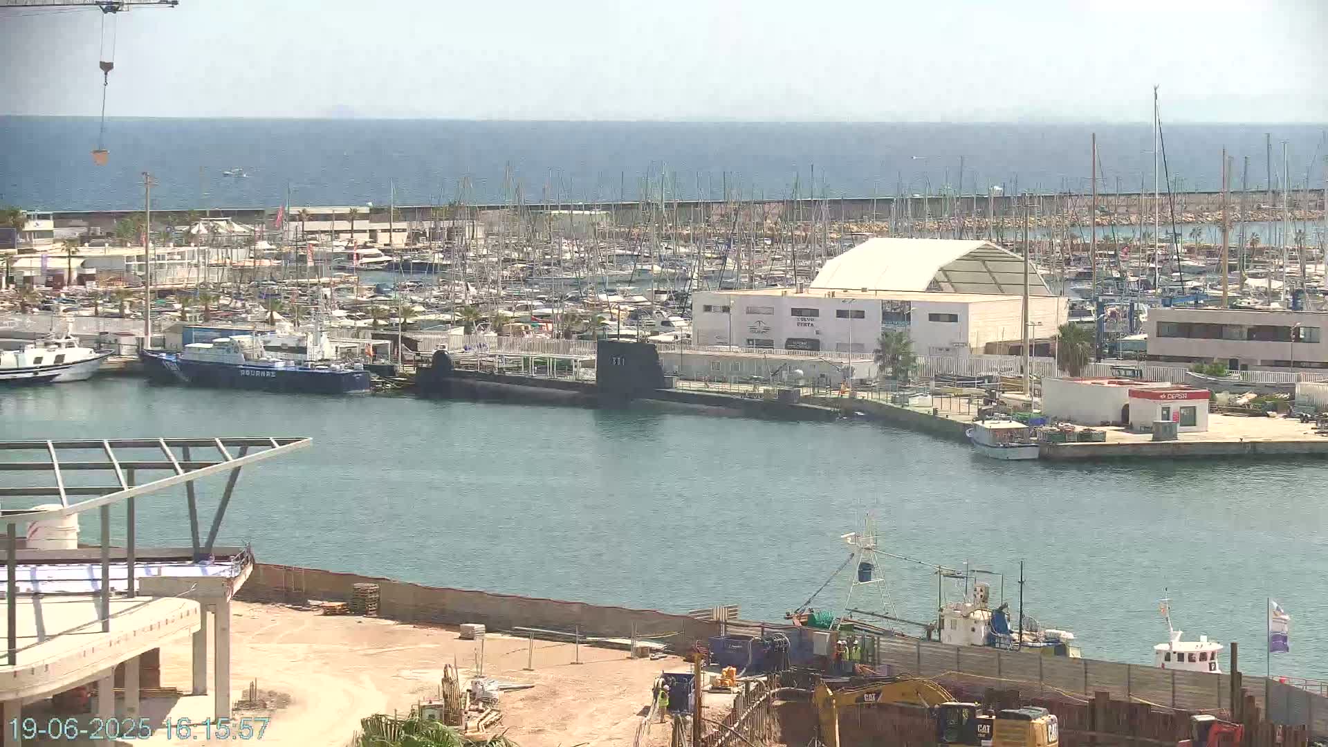 A harbor filled with numerous boats and yachts under a sunny sky, features a docked submarine near a building under construction.