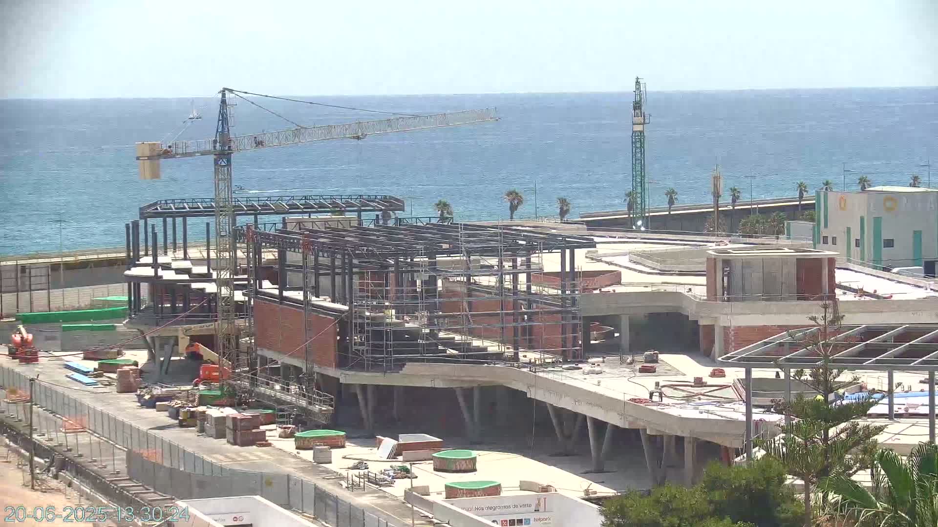 A large building under construction near the ocean on a sunny day, with a crane visible on the site.