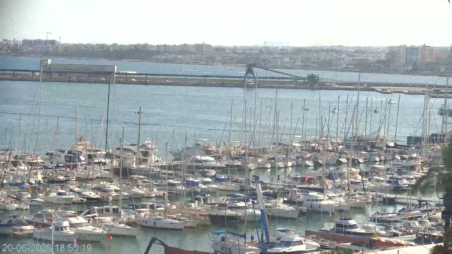 A large marina filled with numerous sailboats and motorboats is visible on a calm, sunny day, with a distant cityscape and industrial structures in the background.
