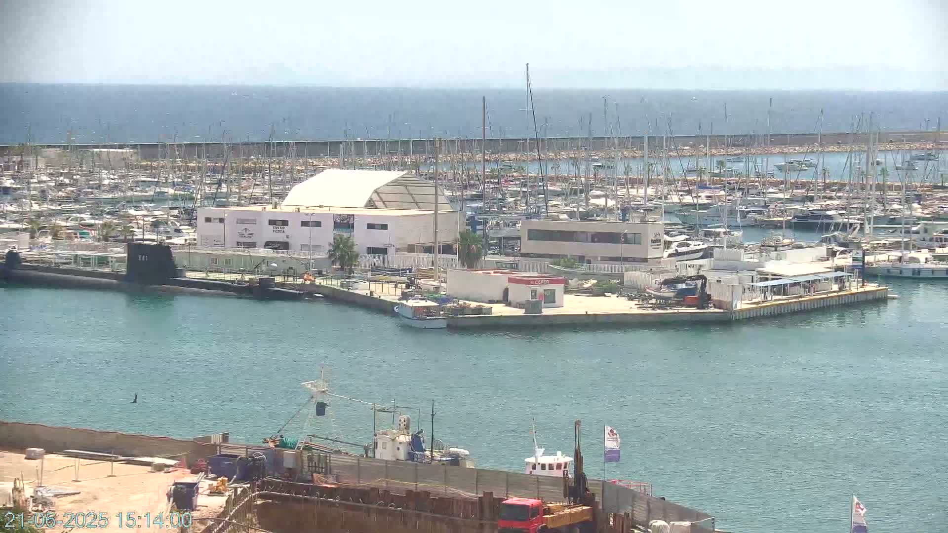 A marina filled with numerous boats and yachts under a clear, sunny sky, with a submarine visible in the foreground.