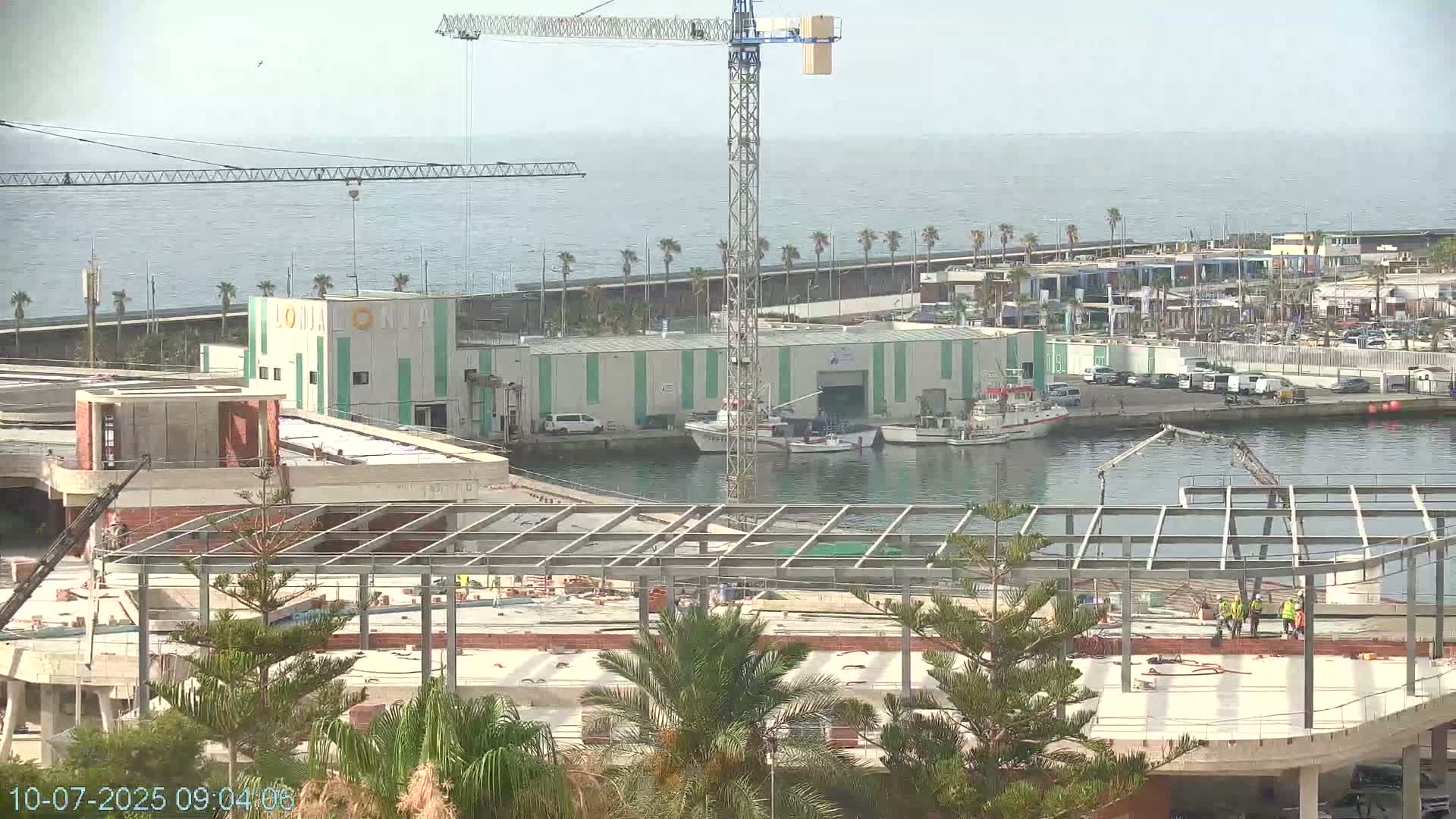 A harbor scene shows a construction site with a metal framework in the foreground, several boats in the water, a building under construction, and a large crane, all under a clear sky.