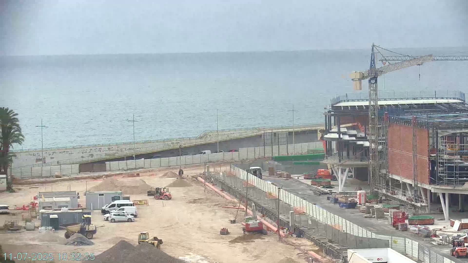 A construction site near a calm ocean under an overcast sky shows heavy machinery, partially built structures, and several vehicles.