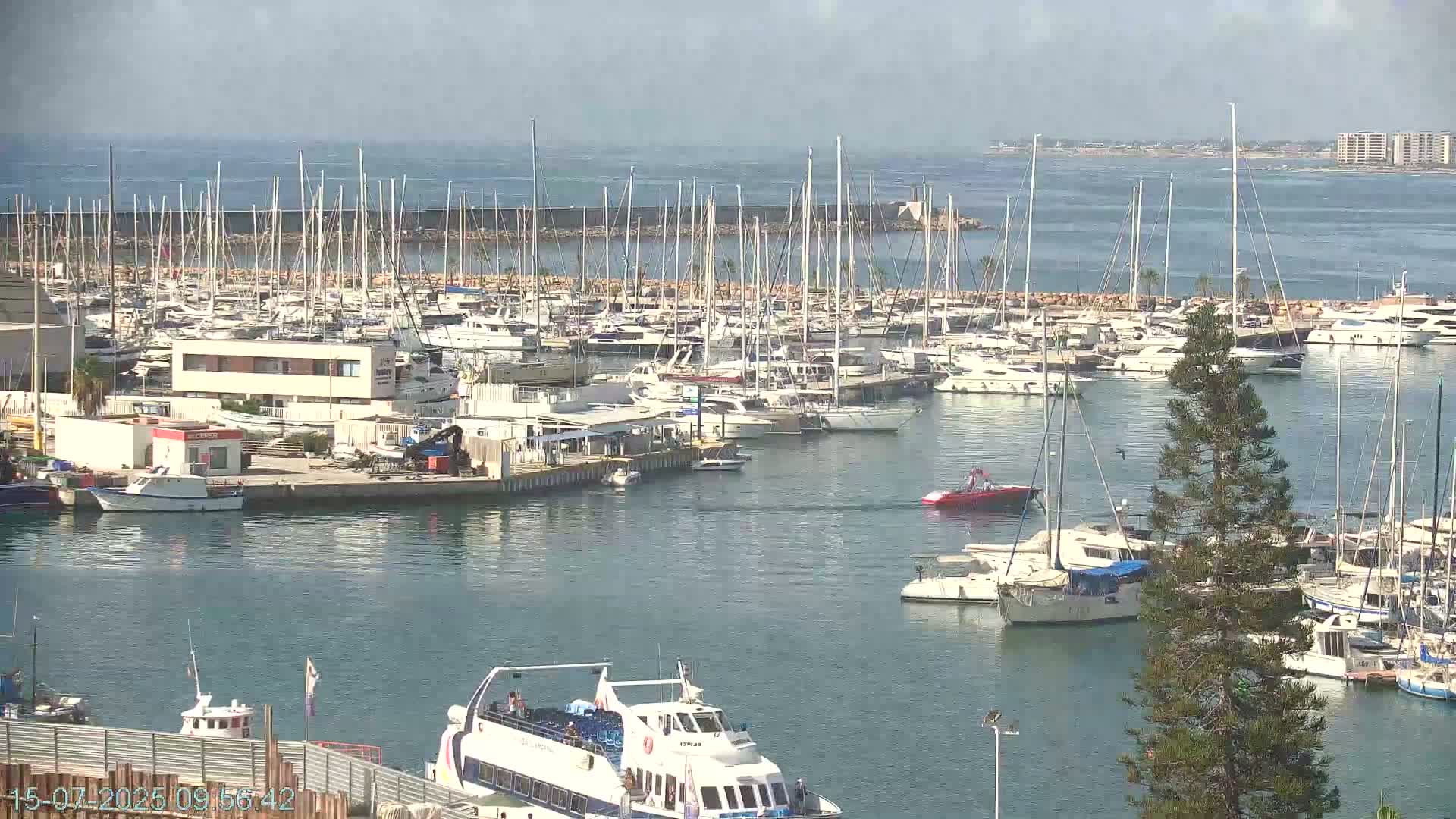 A marina filled with numerous sailboats and motorboats on a partly cloudy day, with calm water and a distant coastline visible.