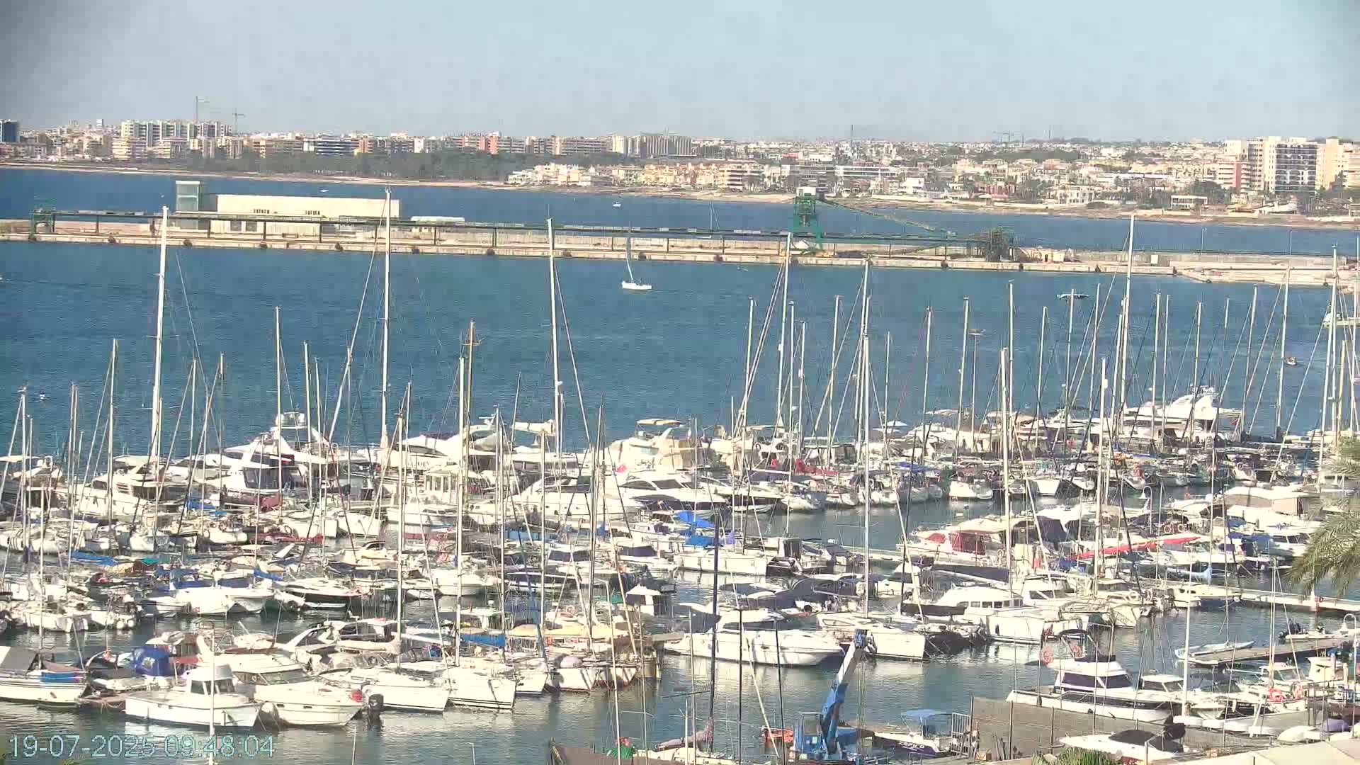 A marina filled with numerous sailboats and motorboats is situated on calm blue water, with a distant cityscape visible under a clear sky.