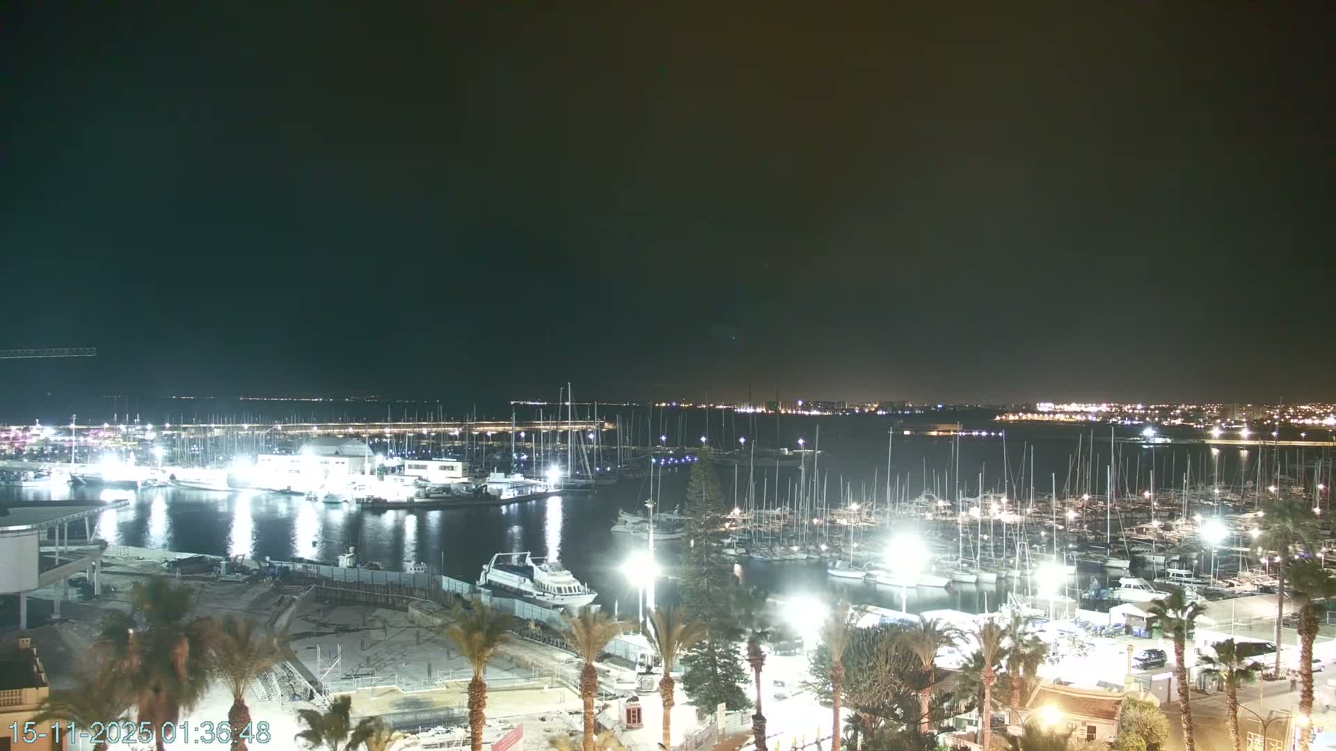 A clear night view overlooks a brightly lit marina filled with numerous sailboats and yachts, with city lights twinkling along the distant coastline.