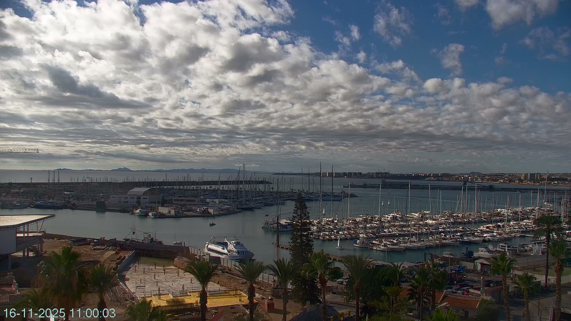 A bustling marina, densely packed with various boats and yachts, is visible under a partly cloudy sky, flanked by coastal development and distant land, with a construction site and palm trees in the foreground.