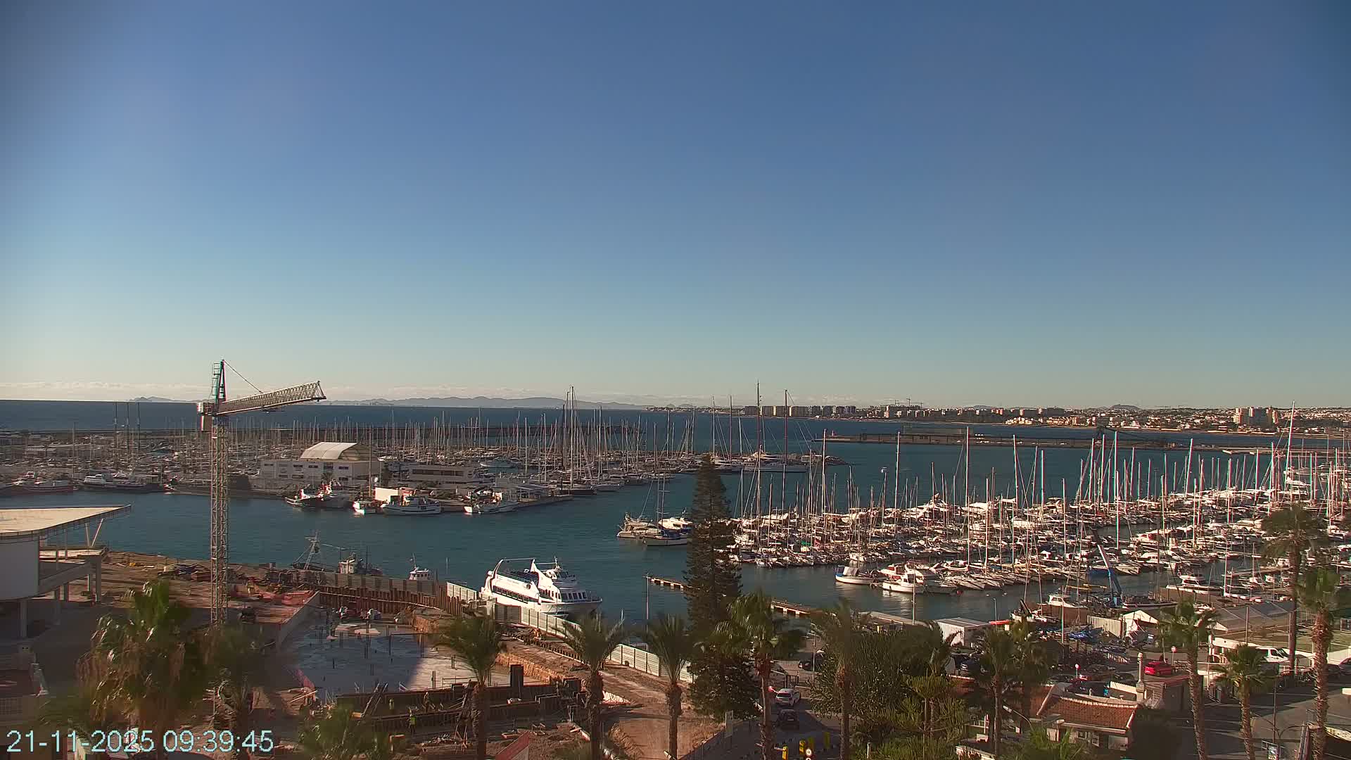 A bustling marina filled with numerous sailboats and yachts is seen under a clear, sunny sky, with a construction site and palm trees in the foreground and a distant cityscape along the horizon.