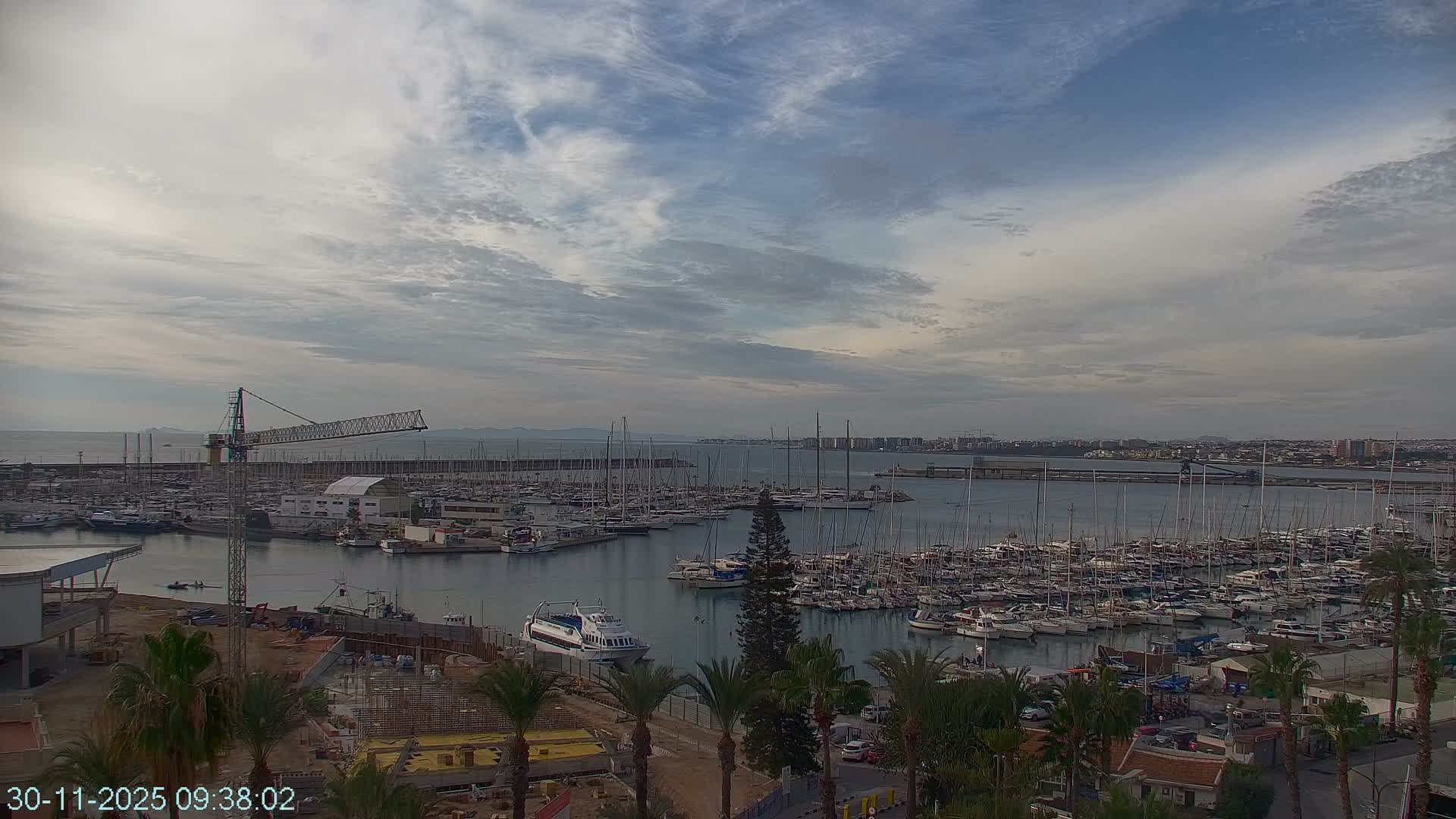An expansive marina teeming with numerous sailboats and motor yachts is seen under a partly cloudy sky, with a construction site and palm trees in the foreground and a cityscape visible in the distance across the water.