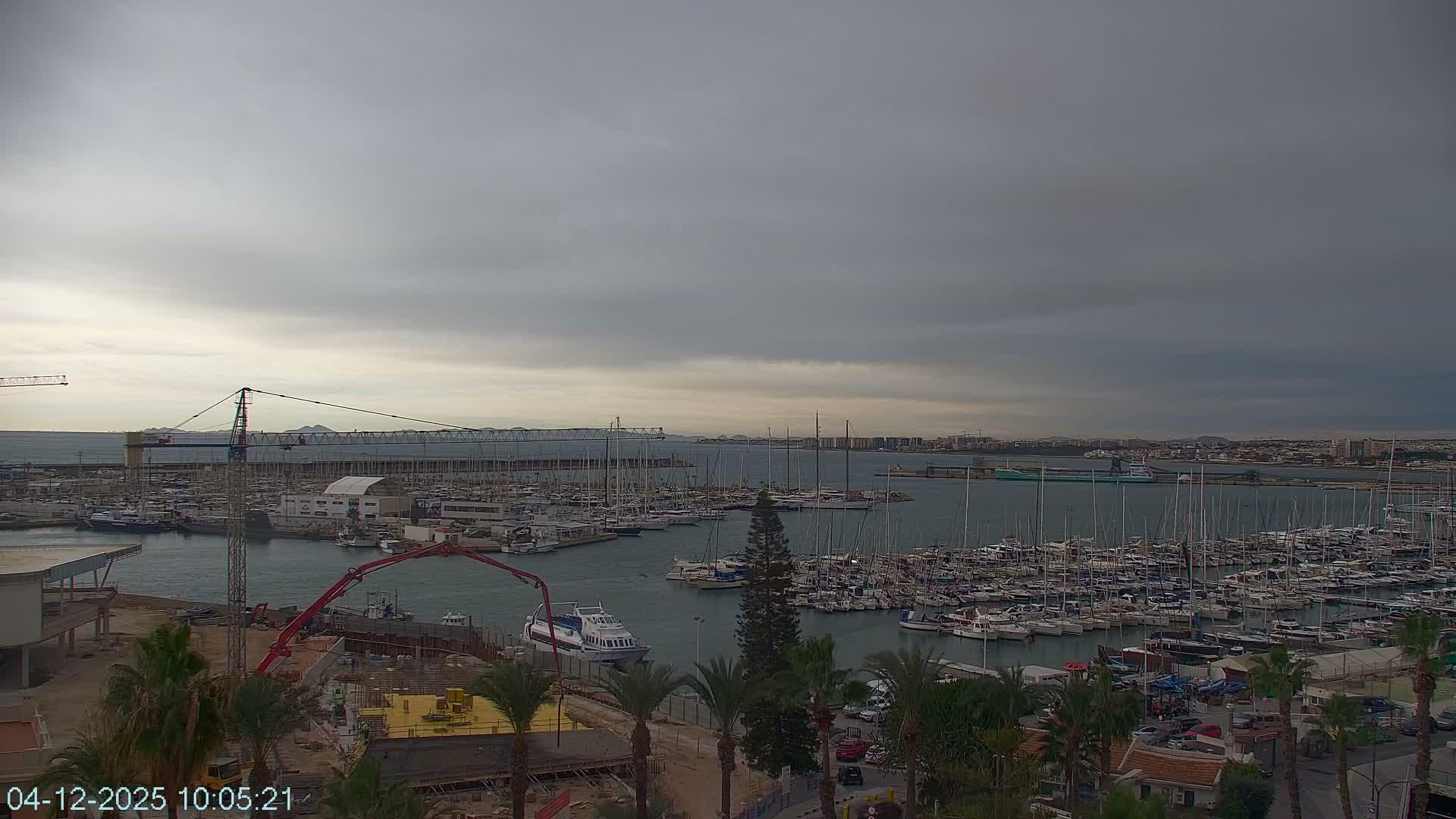 A vast marina filled with numerous boats and yachts is visible under an overcast, cloudy sky, with coastal buildings and active construction in the foreground.