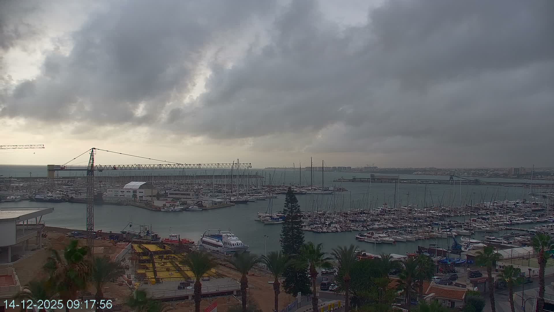 A vast marina filled with numerous boats and yachts is visible under an overcast, cloudy sky, with coastal buildings and active construction in the foreground.