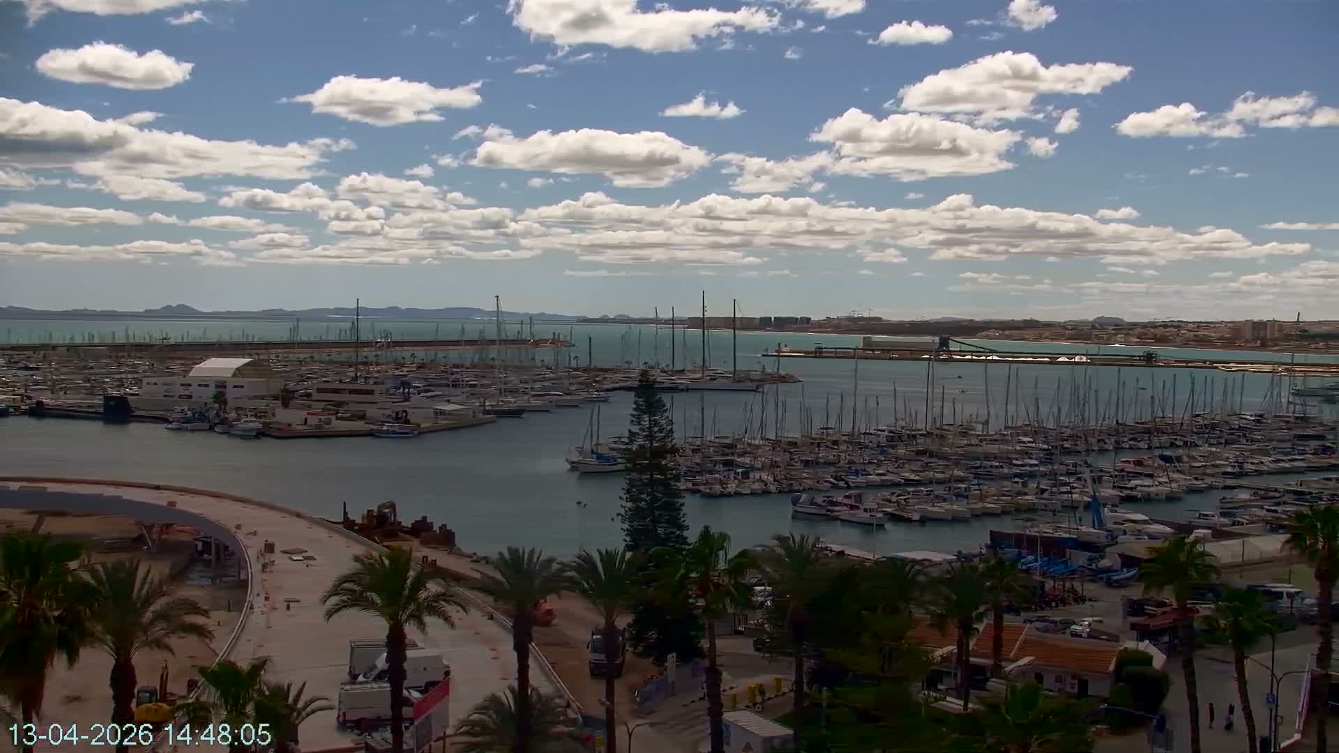 A vast marina filled with numerous boats and yachts is visible under an overcast, cloudy sky, with coastal buildings and active construction in the foreground.
