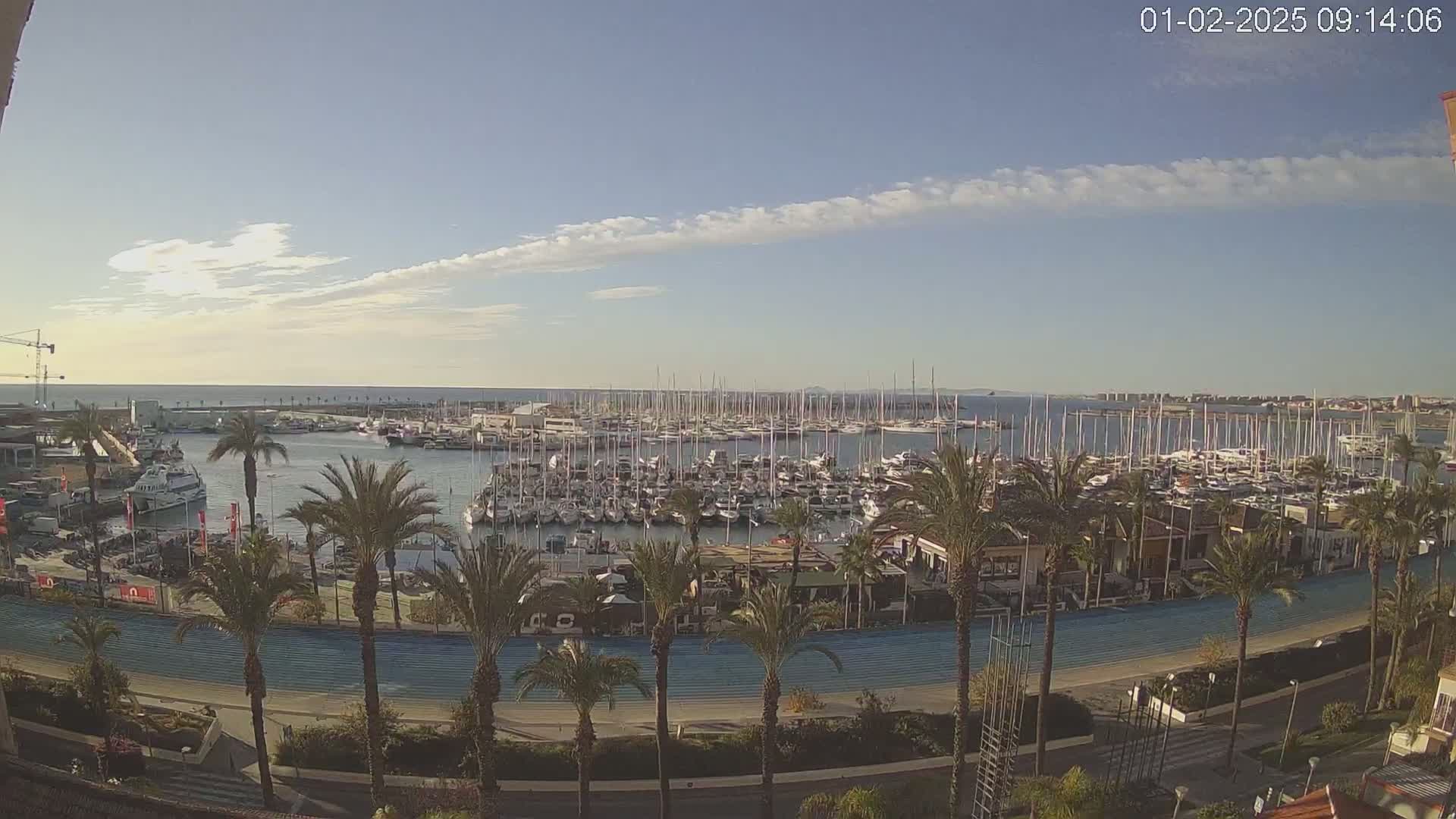 A marina filled with numerous sailboats and motorboats is seen from an elevated vantage point on a partly sunny day with a few thin clouds.