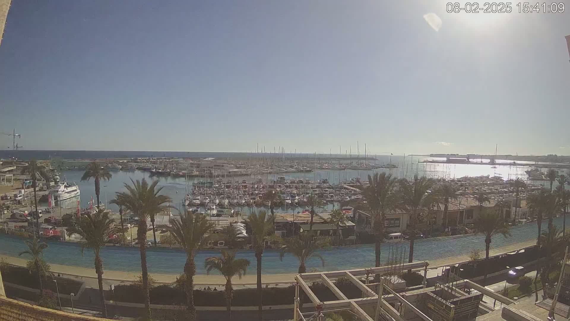 A sunny day overlooking a marina filled with numerous boats, palm trees lining a waterfront promenade, and a partially constructed building in the foreground.