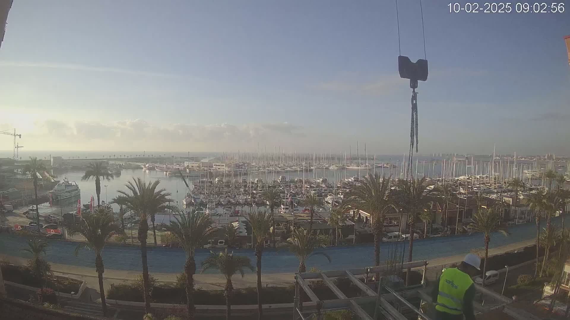 A partly cloudy morning reveals a marina filled with numerous boats and yachts, bordered by palm trees and a waterfront path, with a construction worker visible in the foreground.