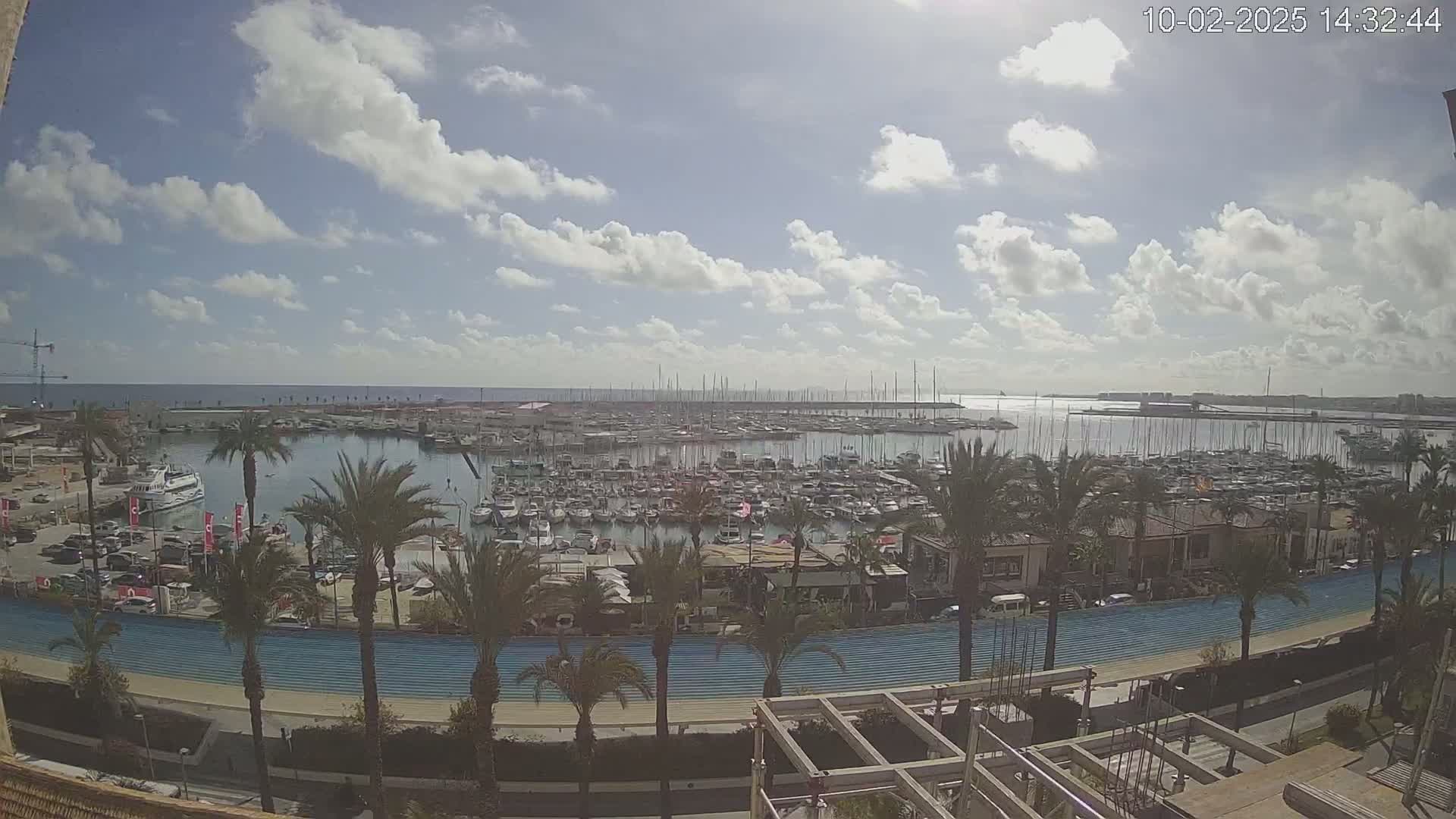 A partly cloudy day overlooks a marina filled with boats, palm trees lining a blue roadway, and a partially constructed building in the foreground.