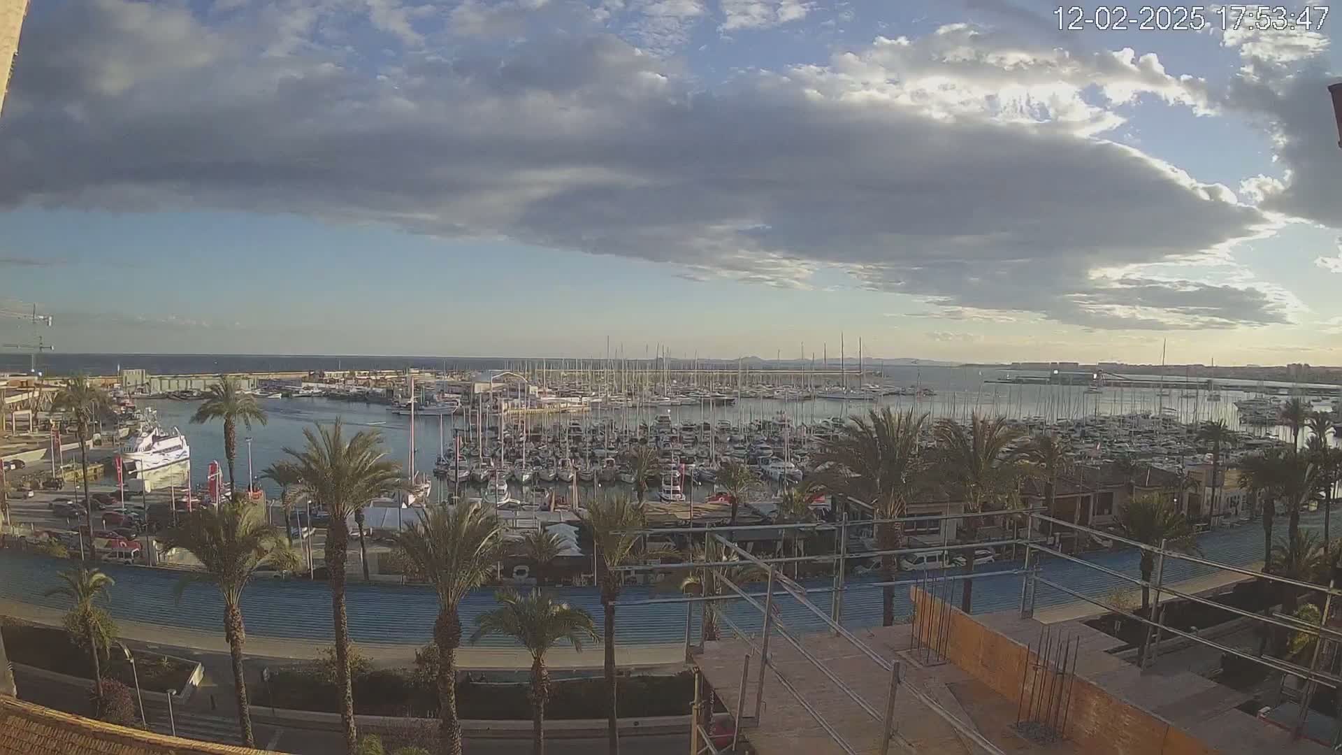 A partly cloudy day overlooks a marina filled with numerous boats and yachts, palm trees lining a roadway next to an under-construction building.