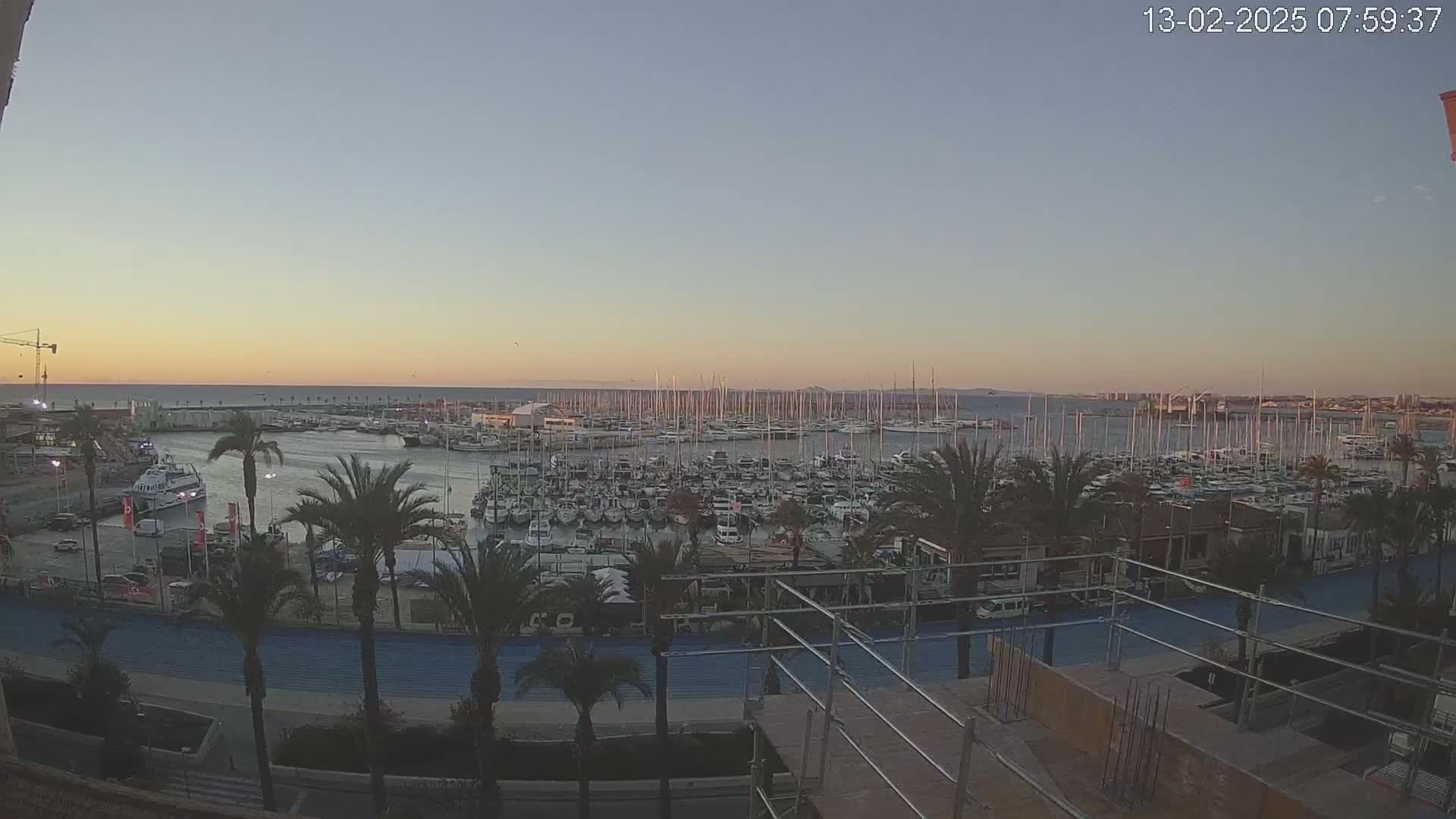 A large marina filled with sailboats and motorboats is seen at sunset, with palm trees lining a waterfront promenade and construction underway in the foreground.
