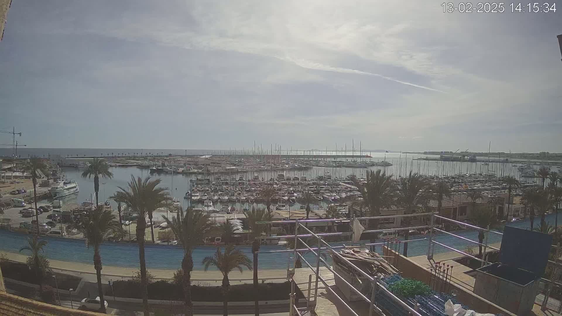 A mostly sunny day reveals a crowded marina filled with sailboats and motorboats, palm trees lining the waterfront, and a nearby roadway under construction.
