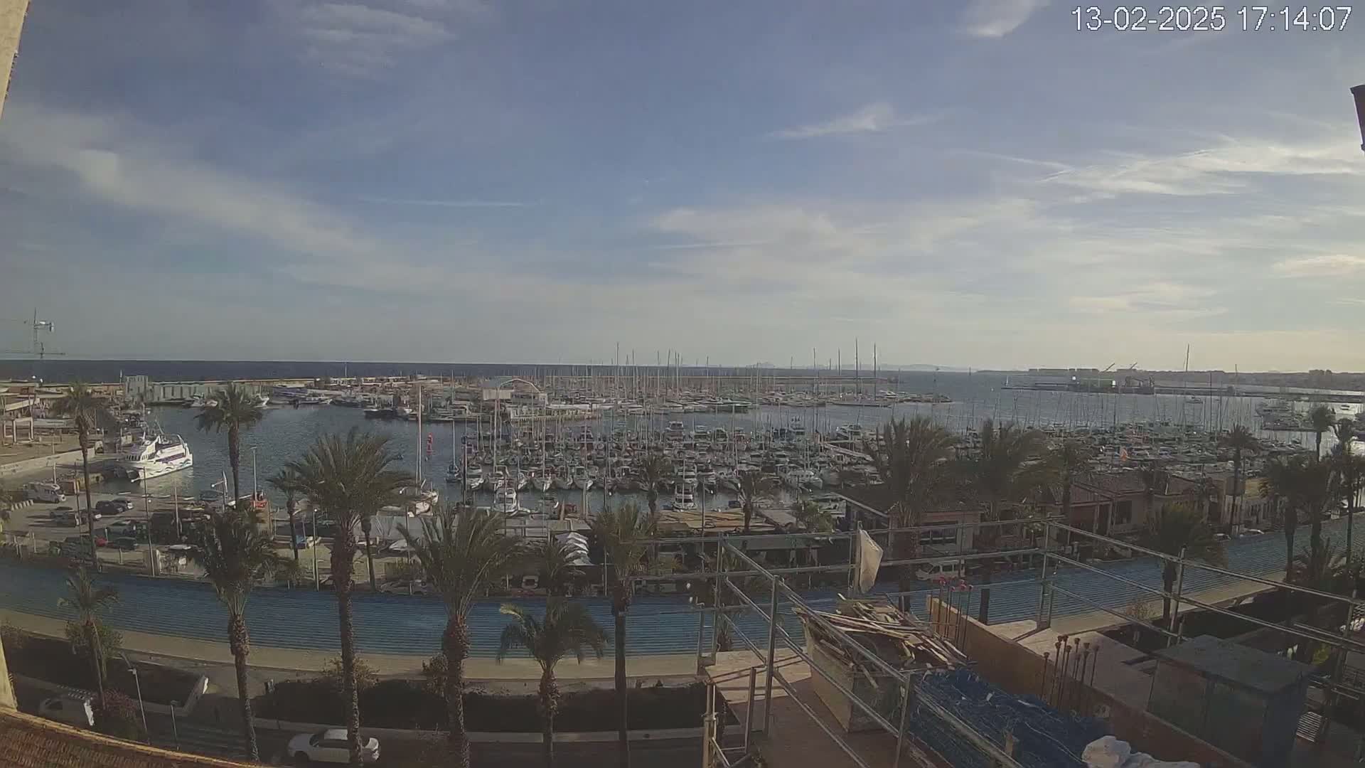 A marina filled with numerous boats and yachts is seen under a partly cloudy sky, surrounded by palm trees and buildings.