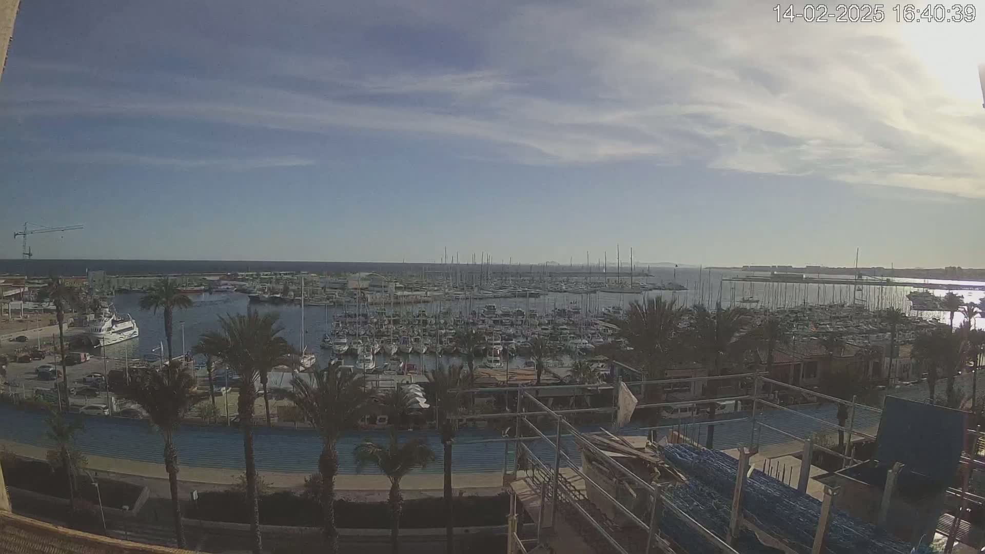 A partly cloudy day reveals a marina filled with numerous boats and sailboats, surrounded by palm trees and a waterfront area with construction underway.