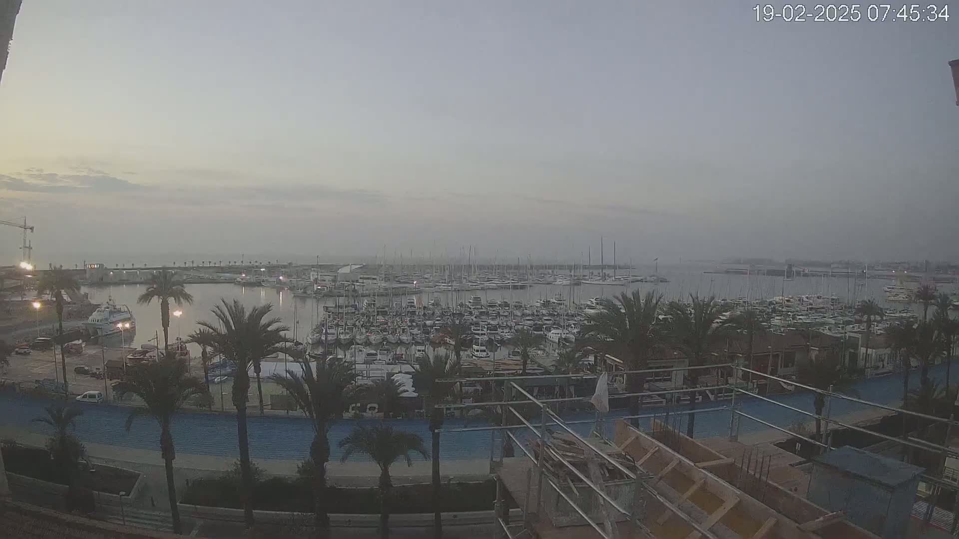 A hazy sunrise over a marina filled with numerous boats, bordered by palm trees and a waterfront walkway under construction.