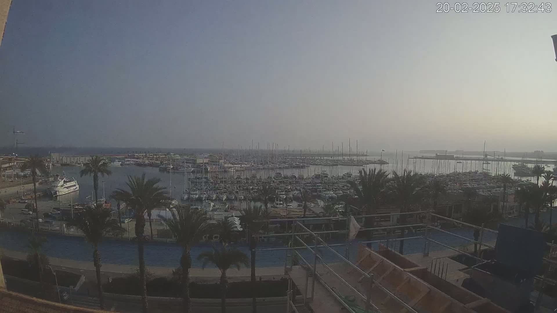A hazy, dusk-lit marina filled with numerous boats and sailboats is viewed from a high vantage point overlooking palm trees and a construction site.