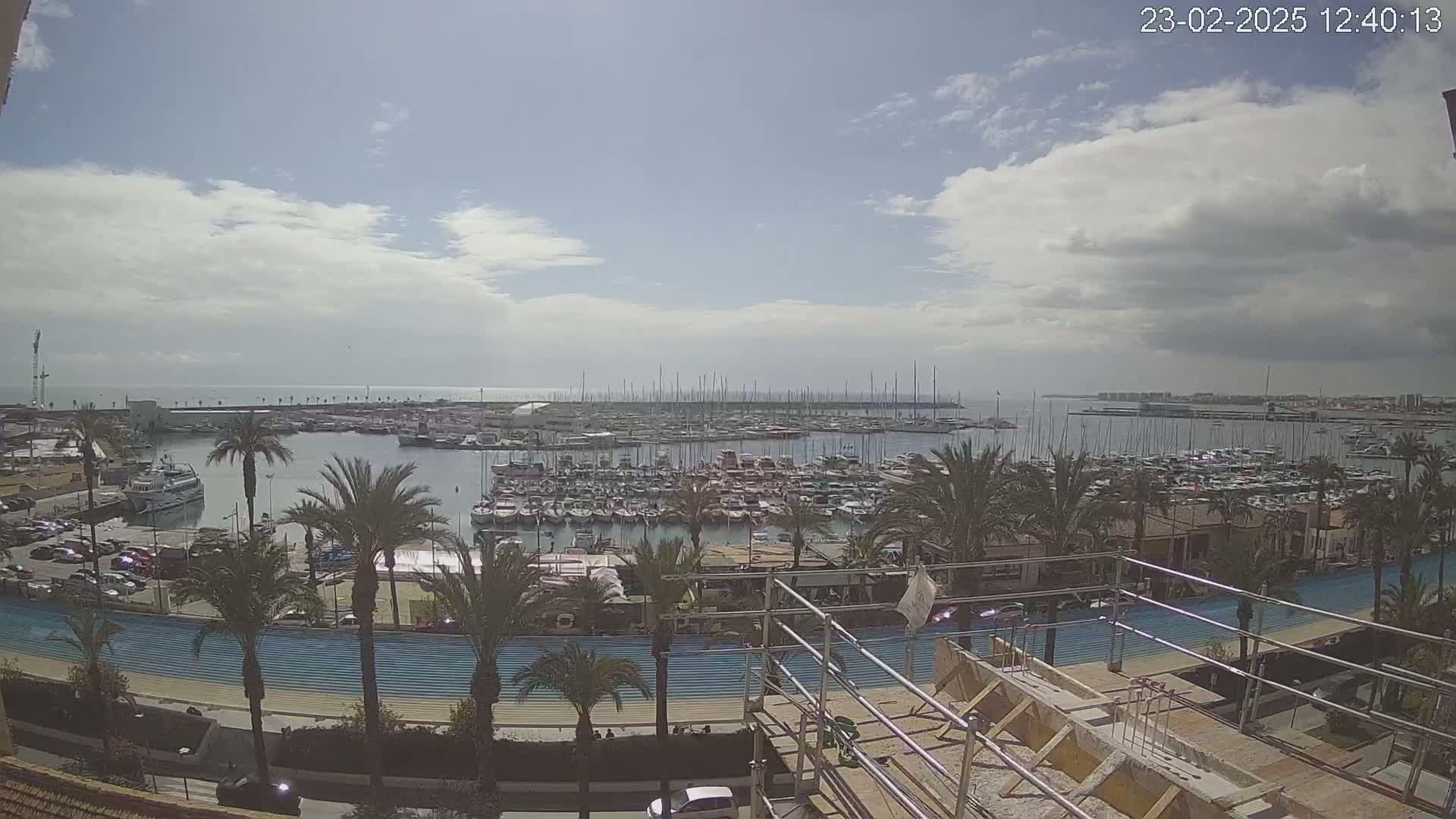 A partly cloudy day overlooks a harbor filled with numerous boats and yachts, surrounded by palm trees and a waterfront promenade with ongoing construction.