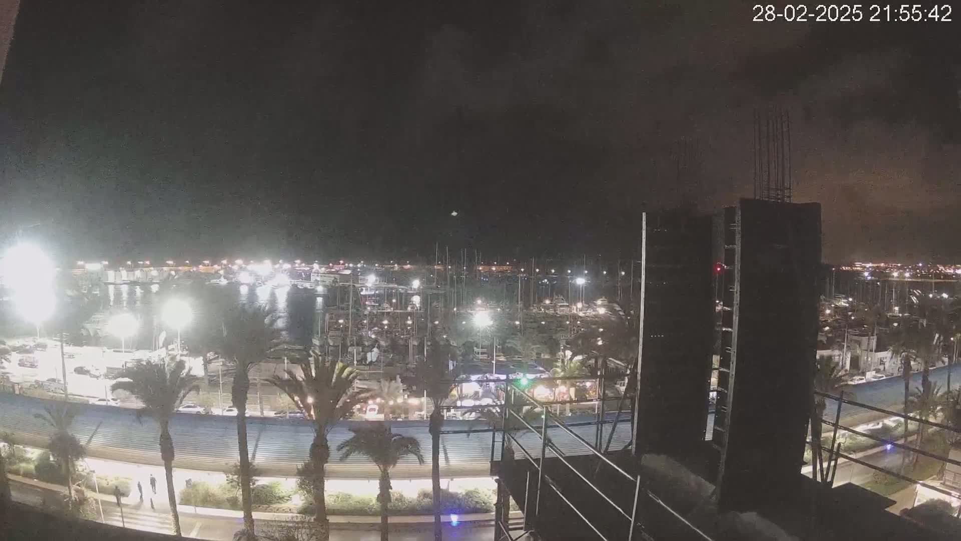 A nighttime view of a marina filled with boats, illuminated by lights, with palm trees and a building under construction visible in the foreground under a partly cloudy sky.