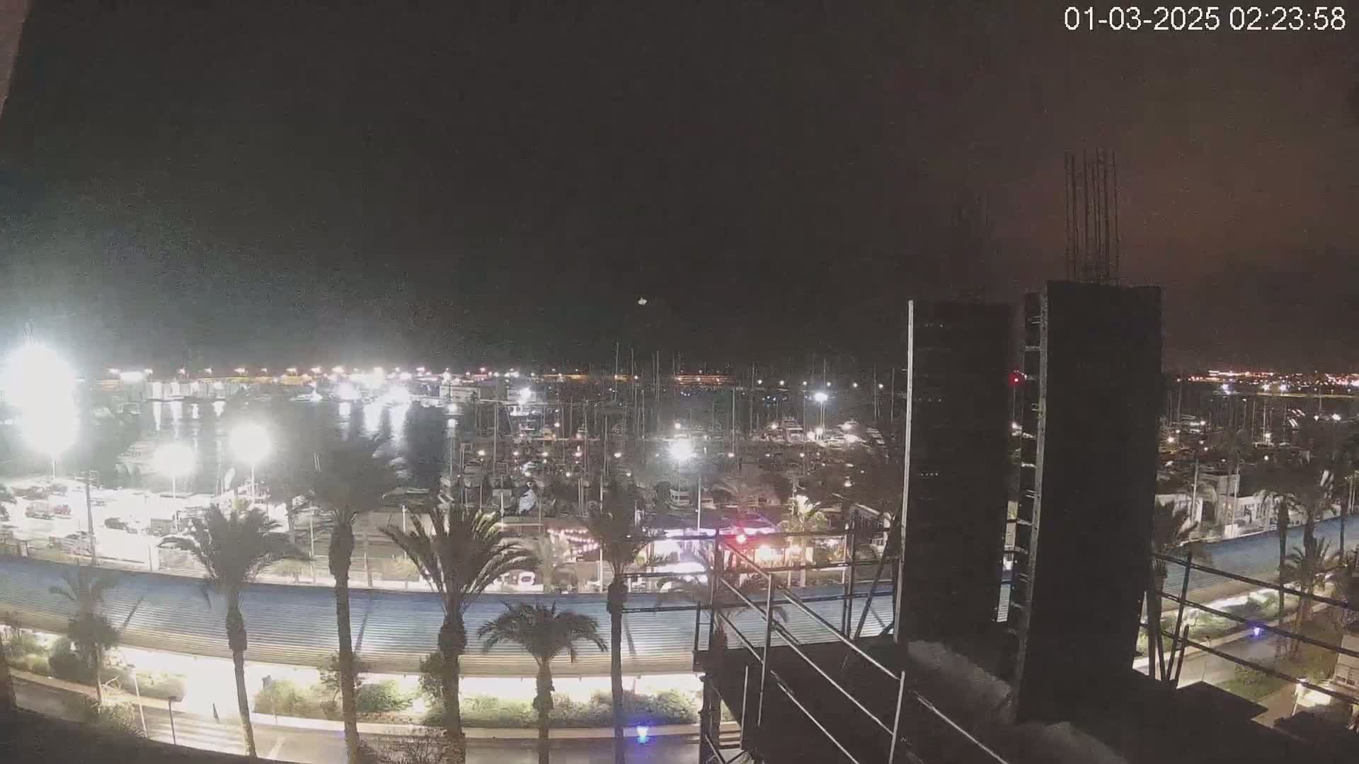 A nighttime view of a marina filled with boats, illuminated by lights, with palm trees lining a nearby roadway and a building under construction in the foreground.
