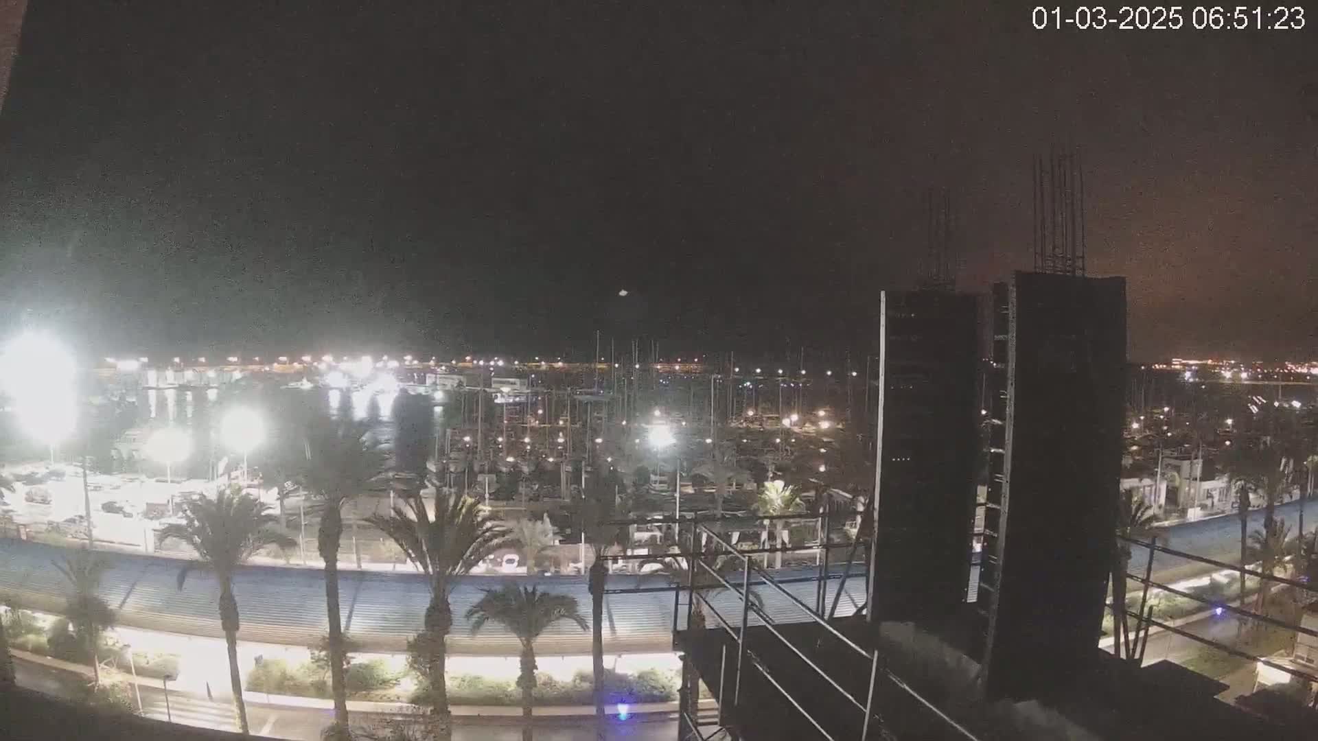 A nighttime view of a marina filled with boats, palm trees lining a roadway, and a building under construction in the foreground.