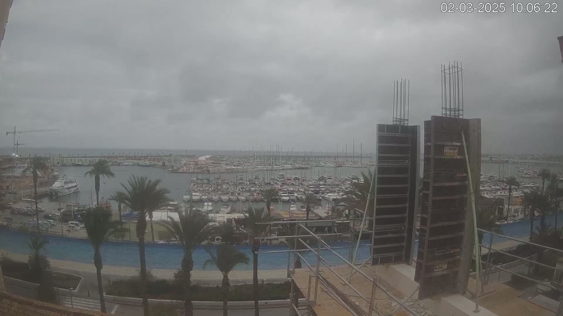 A marina filled with numerous boats is viewed from a construction site under an overcast sky.