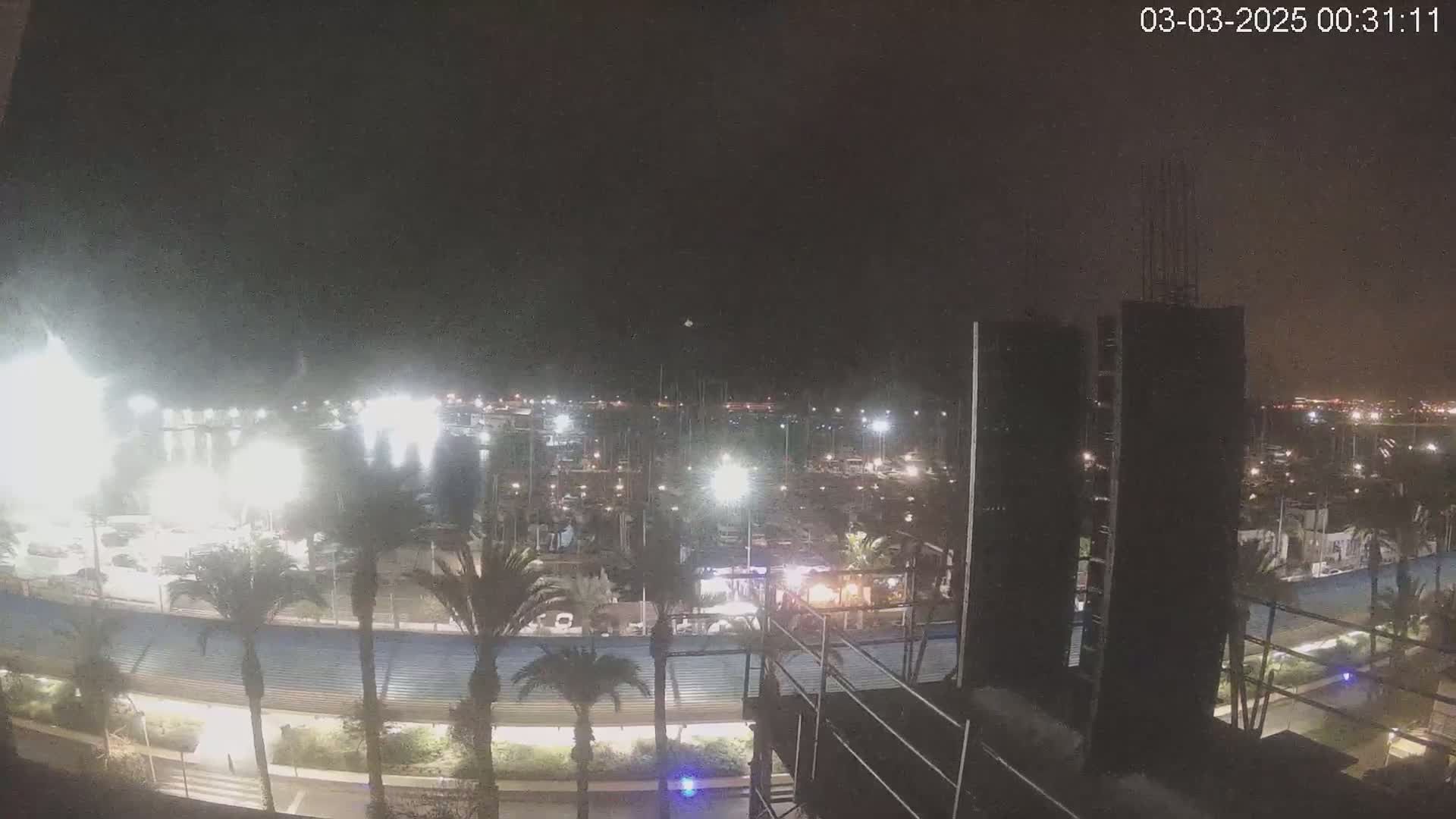 A nighttime view of a marina filled with boats, illuminated by lights, with palm trees lining a nearby walkway and a building under construction in the foreground.