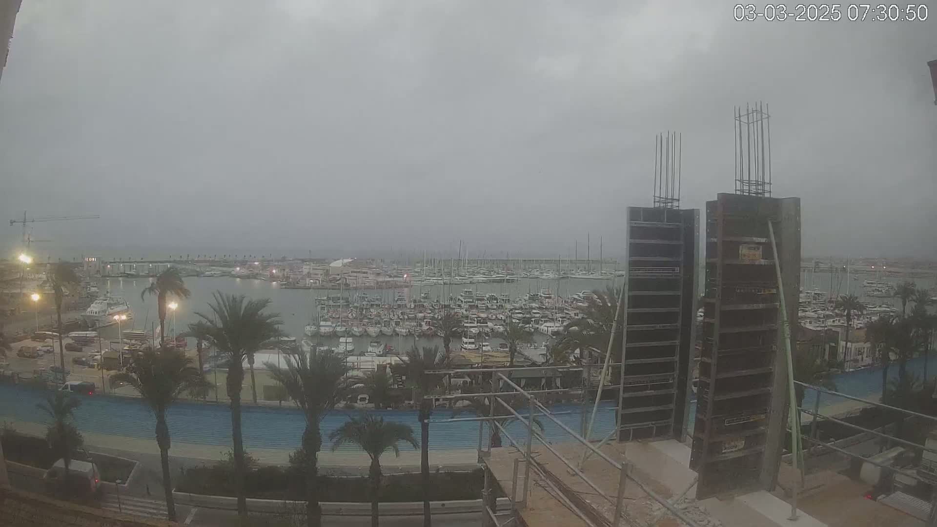 A marina filled with numerous boats is viewed from a distance under an overcast sky, with construction underway in the foreground.