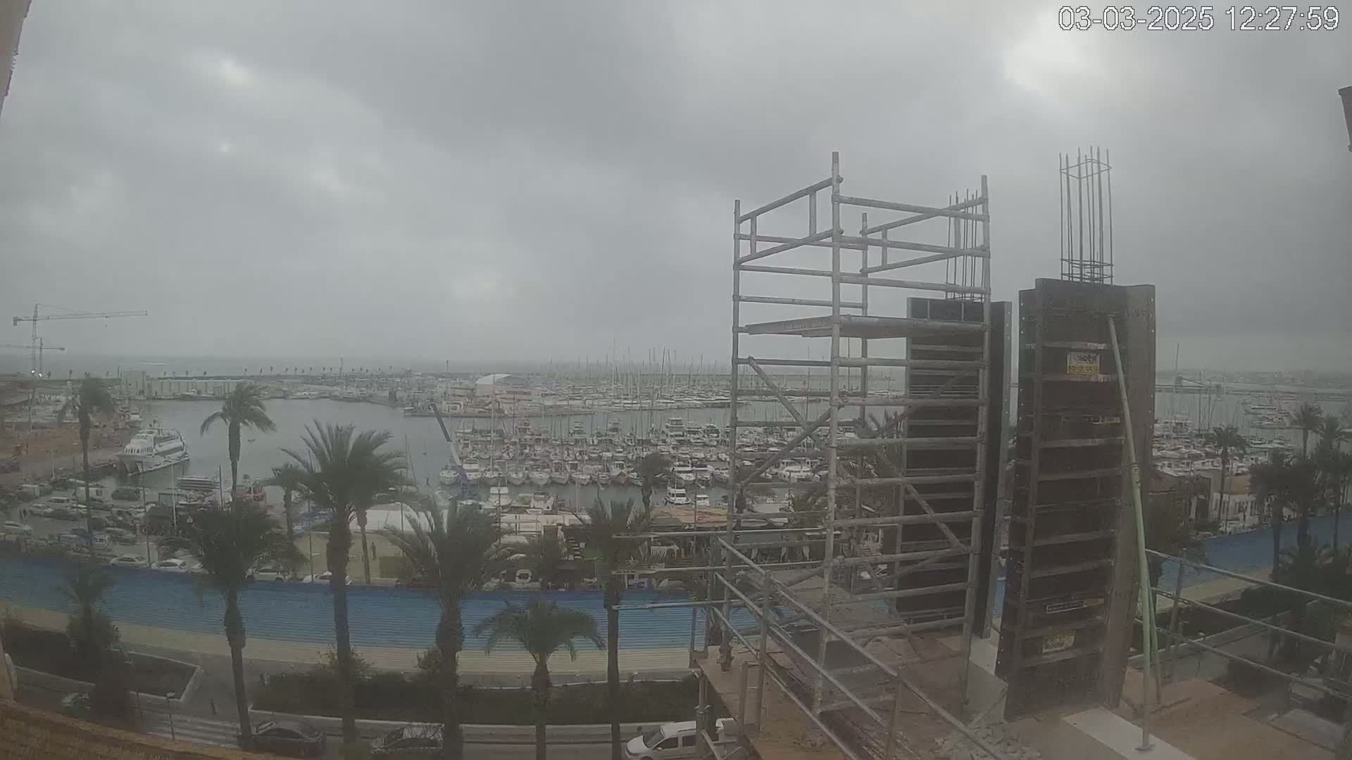 A construction site with scaffolding overlooks a marina filled with boats on an overcast day.