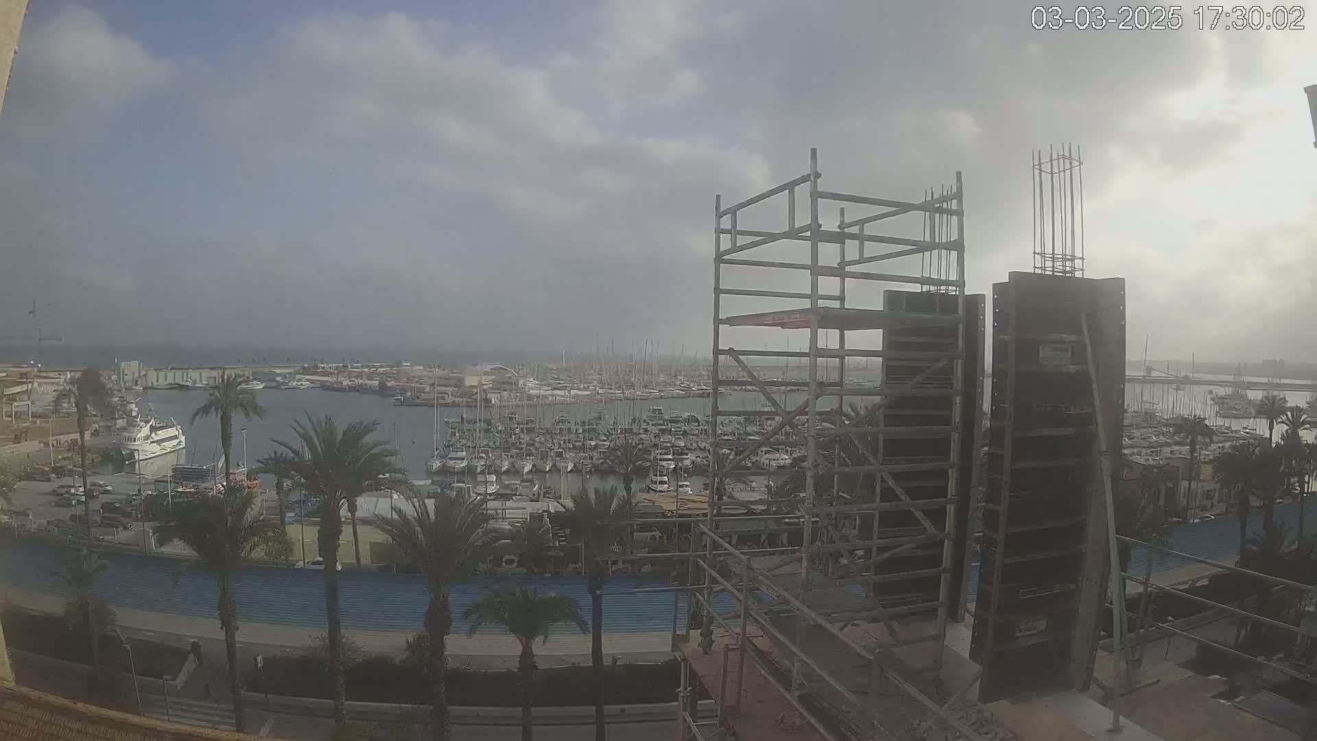 A view from a construction site shows a marina filled with boats under a cloudy sky.