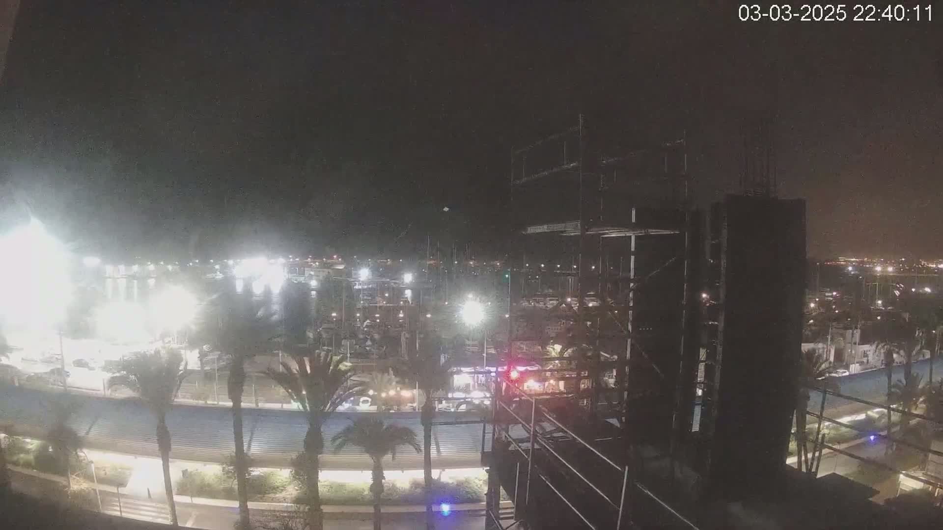 A nighttime view of a marina and palm trees, with a building under construction in the foreground, under clear skies.