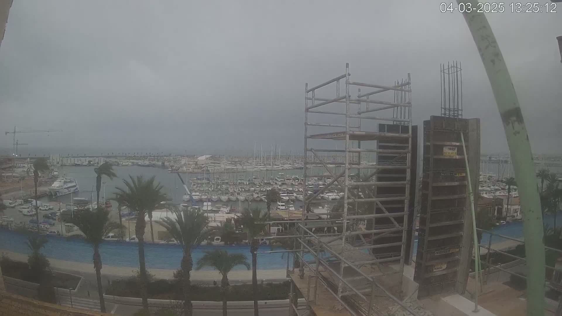 A construction site with scaffolding overlooks a harbor filled with numerous boats under an overcast sky.