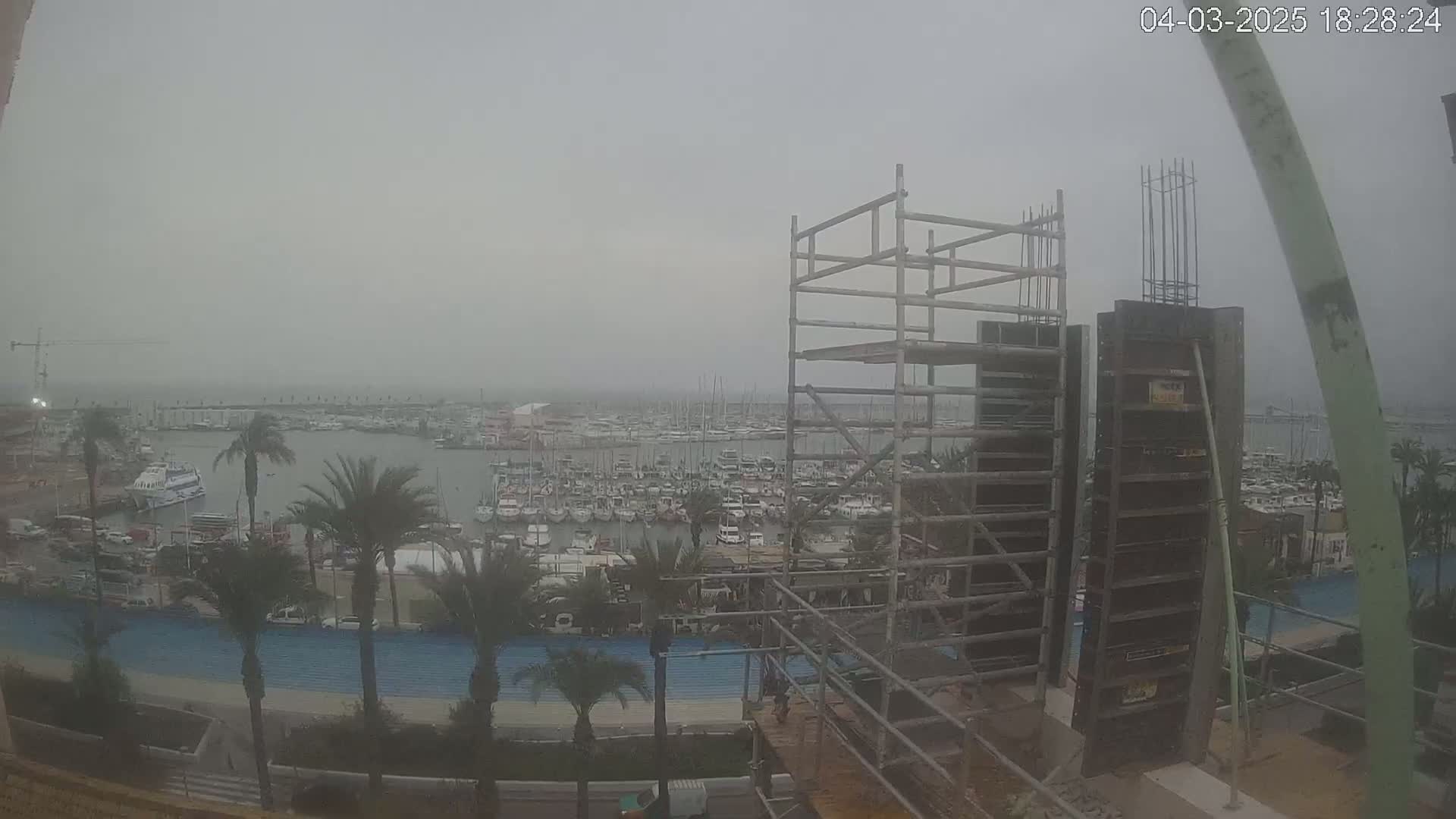 A hazy view of a marina filled with boats is seen from a construction site featuring scaffolding and concrete forms, under an overcast sky.