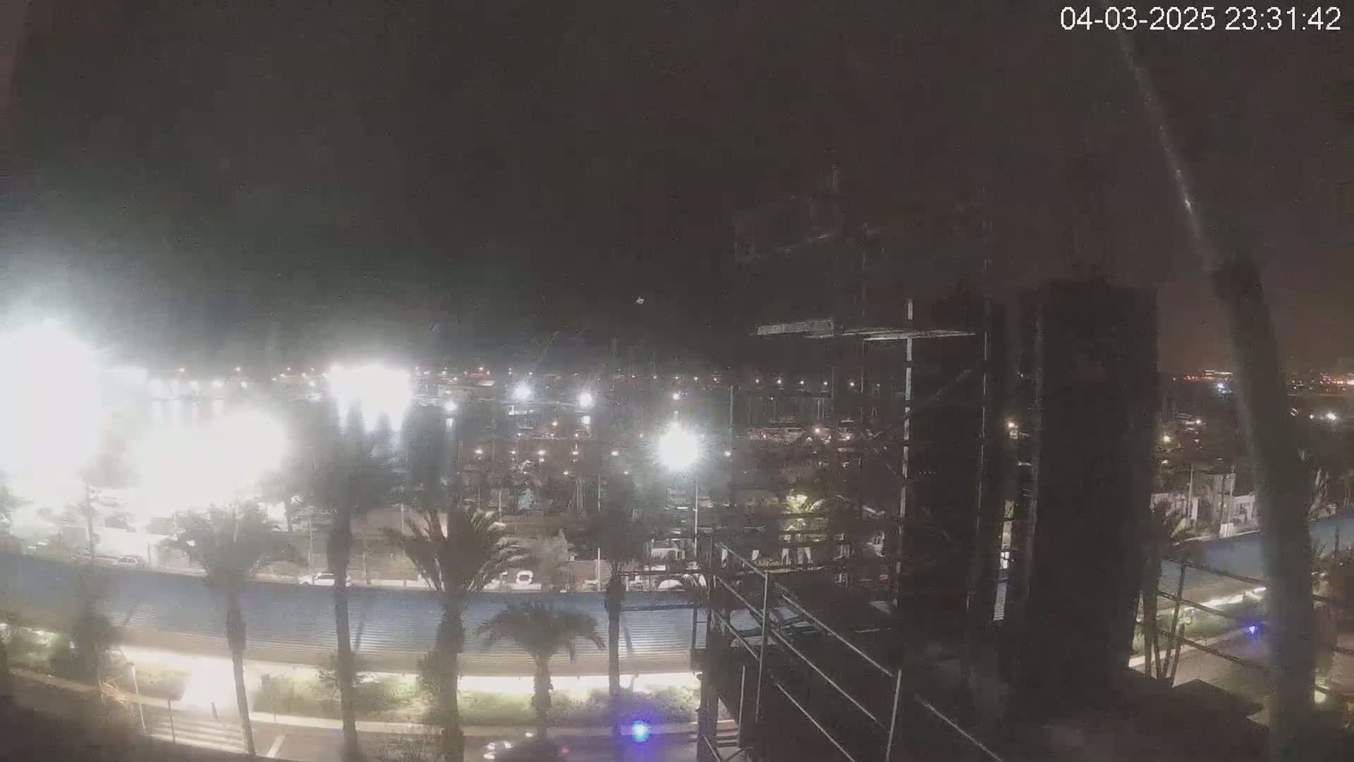 A nighttime view of a marina with bright lights illuminating boats and palm trees, under a dark sky.