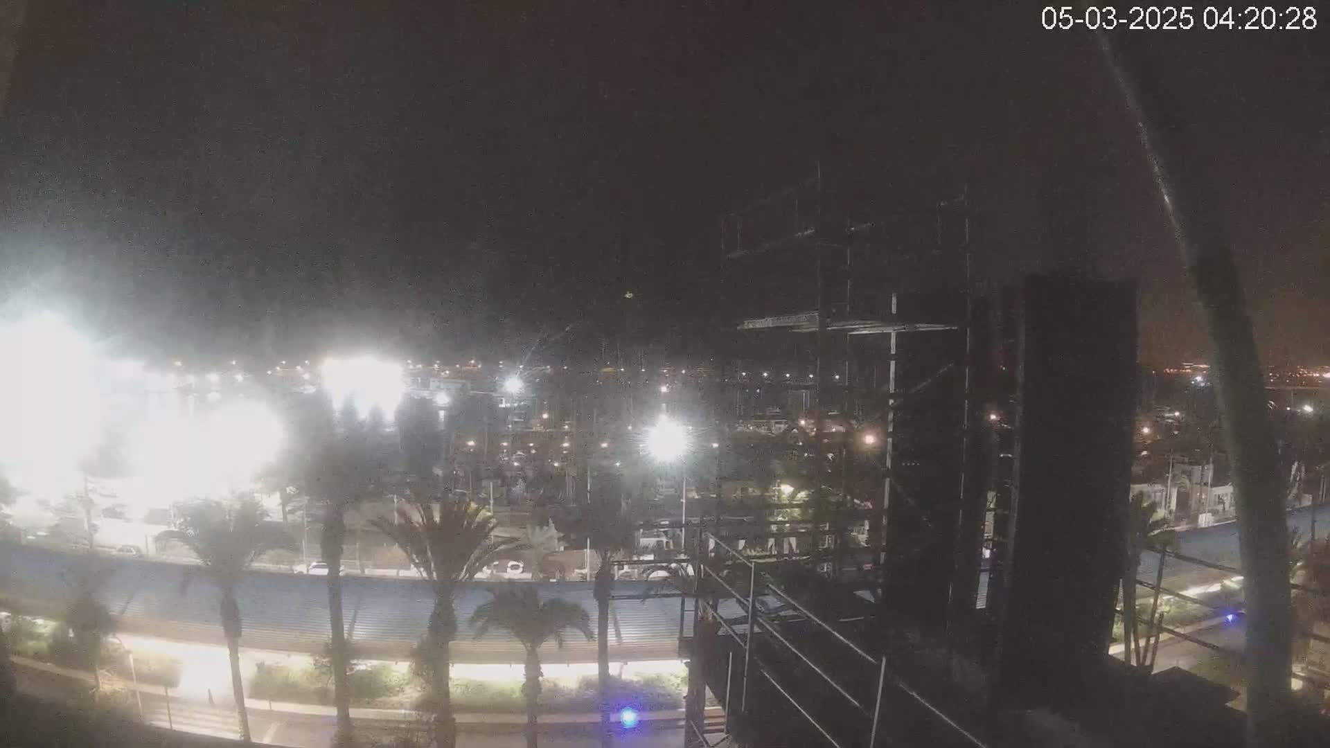 A nighttime, brightly lit marina and stadium are visible from an elevated vantage point, under a dark, possibly cloudy sky.