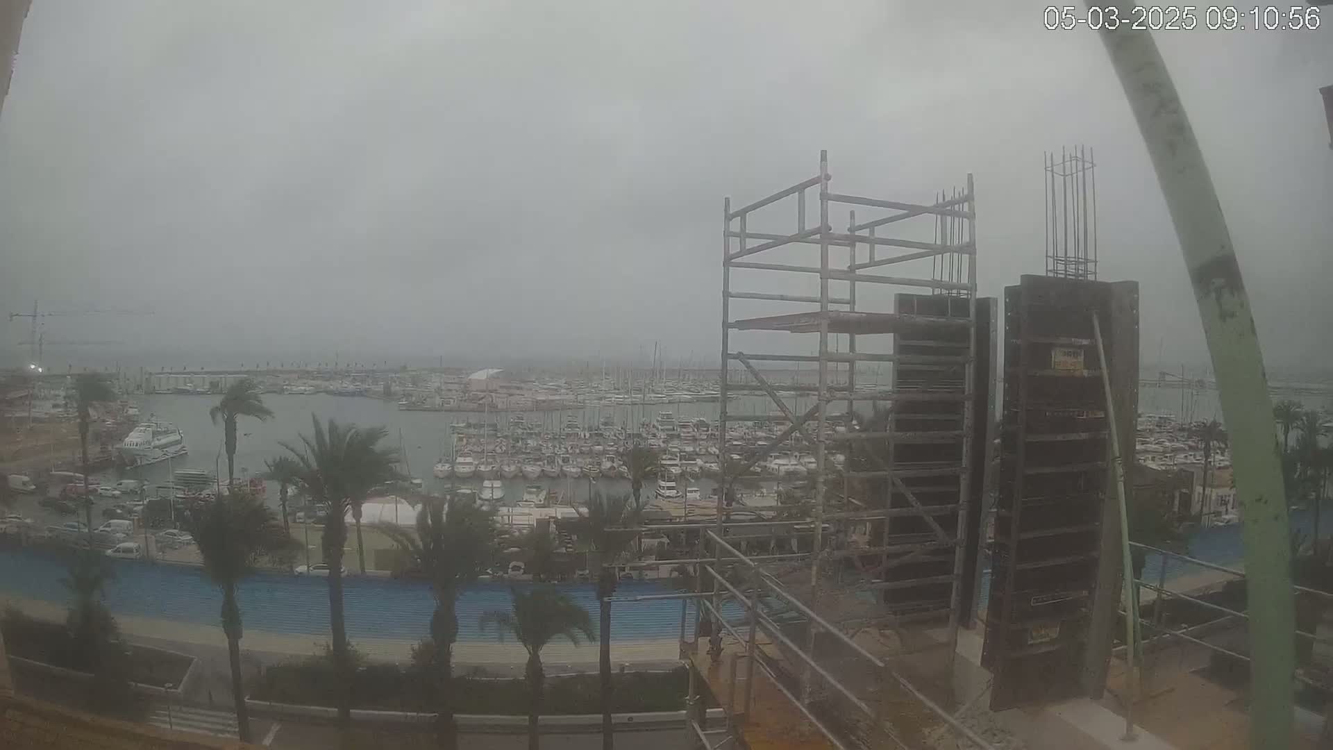 A hazy view from a construction site shows a marina filled with boats and palm trees lining a roadway under a gray sky.
