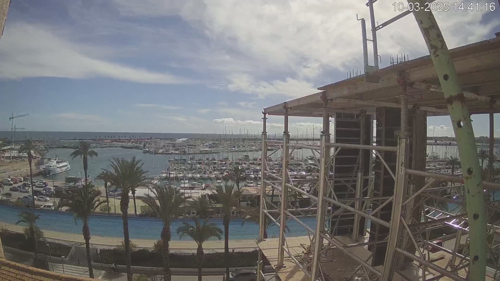 A partly cloudy sky overlooks a marina filled with boats, a waterfront promenade with palm trees, and a building under construction.
