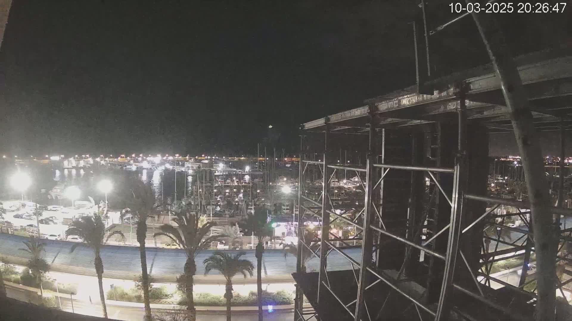 A nighttime view of a marina filled with boats, surrounded by palm trees and a roadway, seen from a vantage point overlooking a metal scaffolding structure.