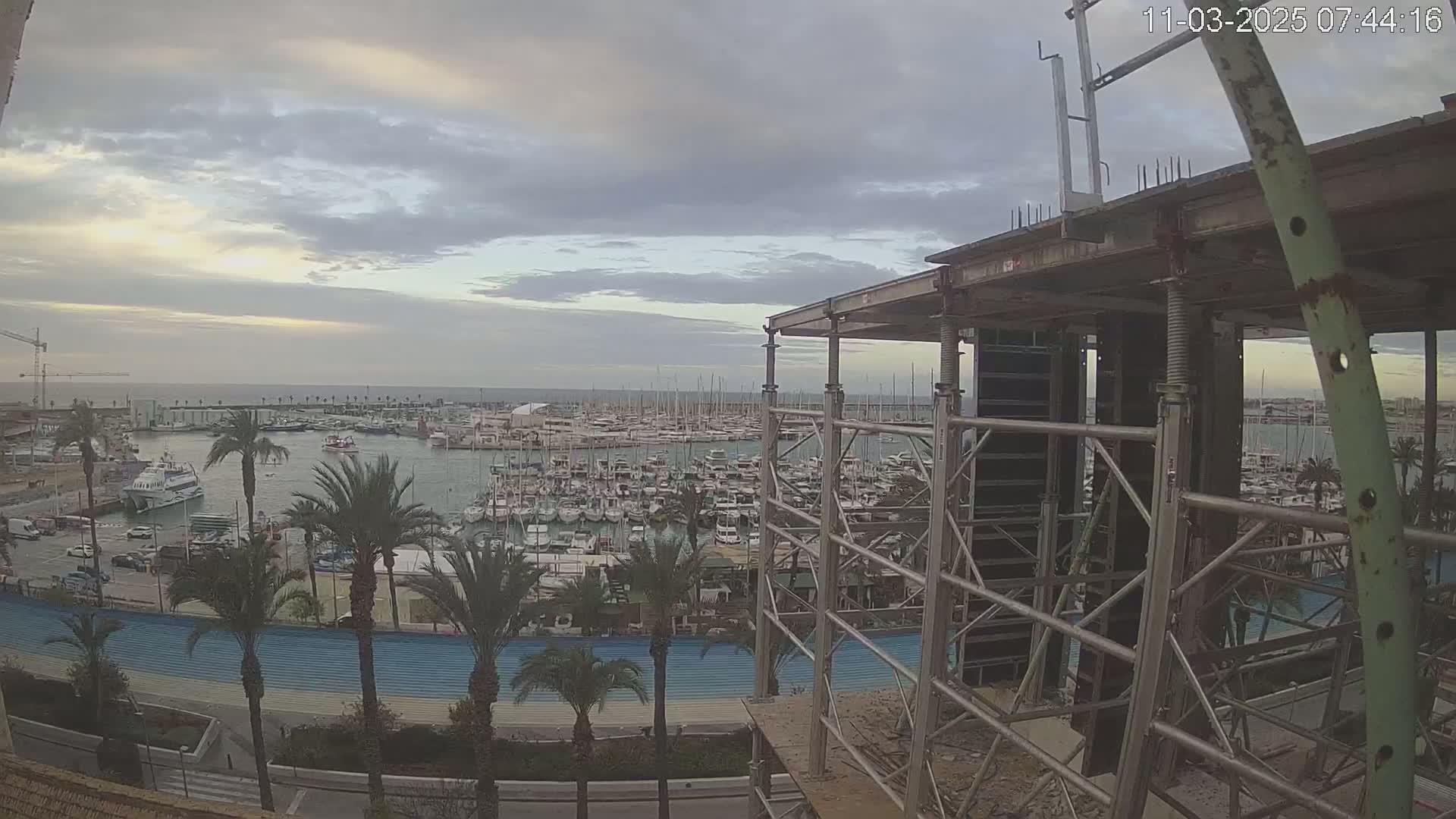 A mostly cloudy sunrise overlooks a harbor filled with many boats and sailboats, seen from a vantage point of a building under construction.
