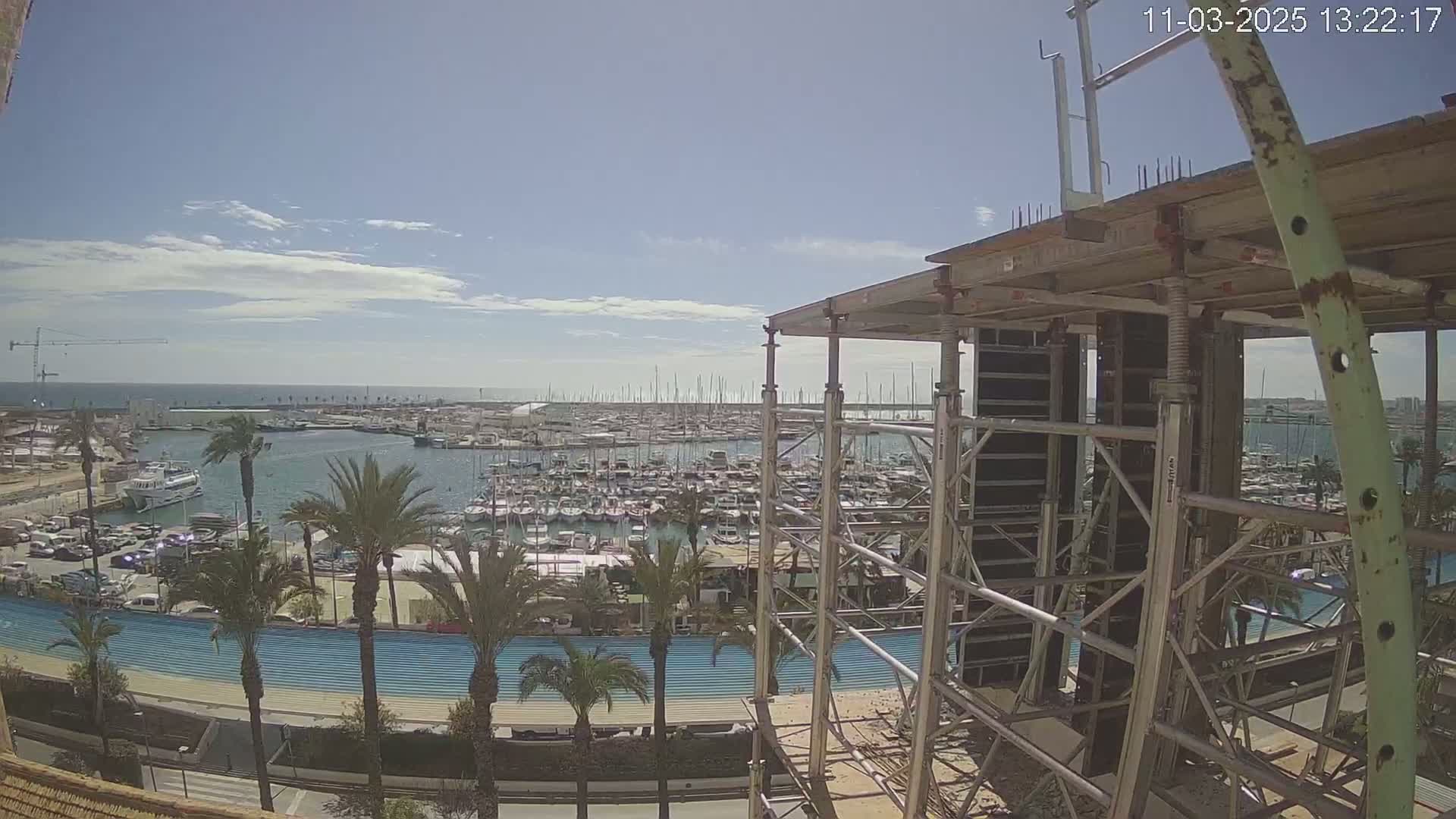 A mostly sunny day shows a view of a marina filled with boats from a partially constructed building, with palm trees and a blue pathway visible in the foreground.