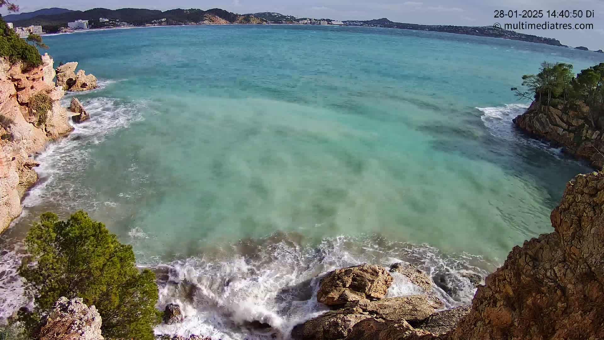 Waves crash against rocky cliffs surrounding a cove of clear, turquoise water, with a town visible in the distance under a partly sunny sky.
